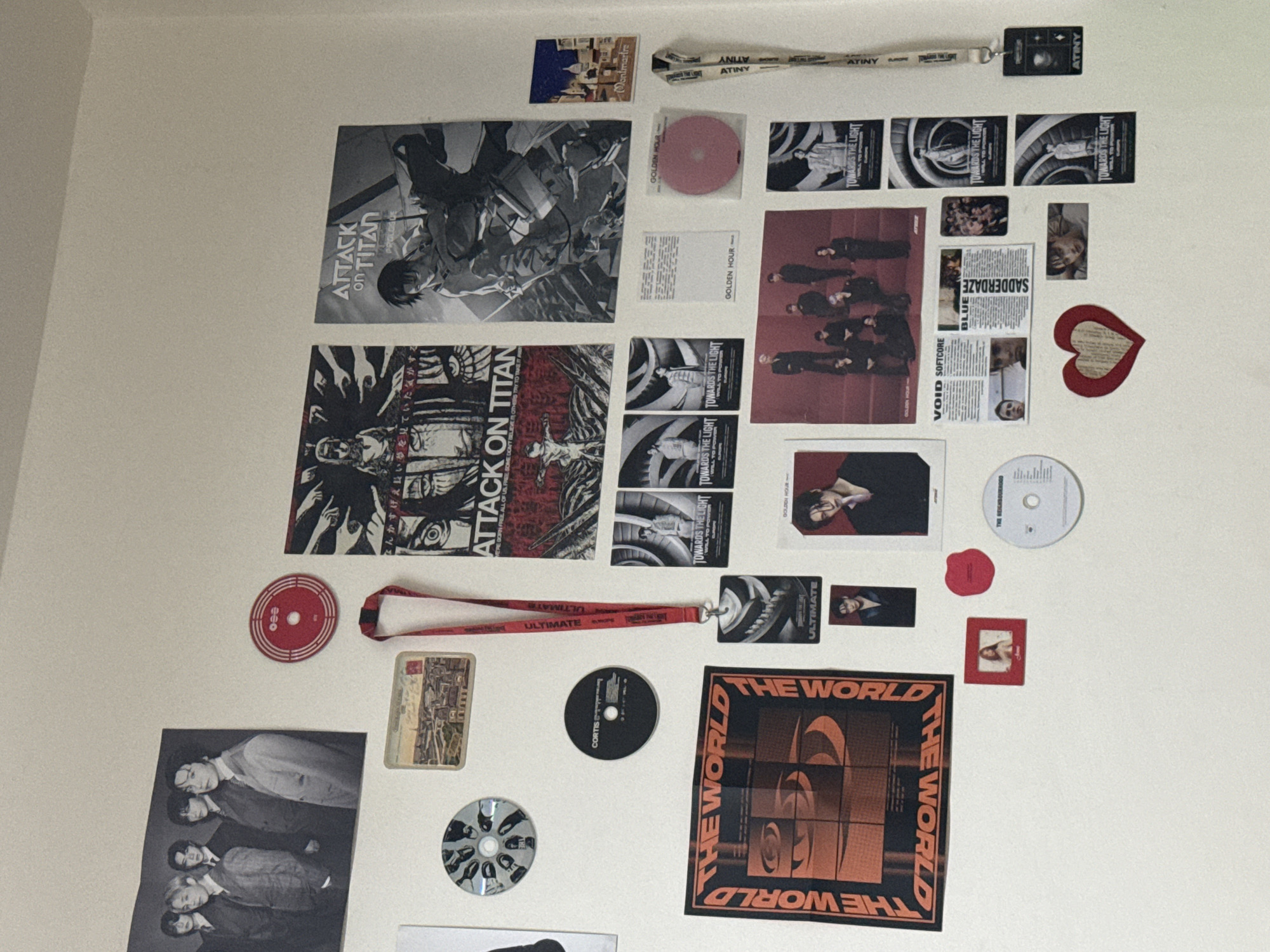 The image shows a collection of posters, cards, CDs, and lanyards attached to a white wall. The items appear to be related to the anime "Attack on Titan" and the K-pop group Ateez. The wall decorations include two large "Attack on Titan" posters, multiple smaller cards and posters of Ateez, several CDs, and two lanyards with "ATINY" and "ULTIMATE" printed on them. There is also a small card that says Montmartre, and one that says "THE REIGHBOURH" It is not possible to determine the location of the scene as Sofia, Bulgaria from the contents of this image.
