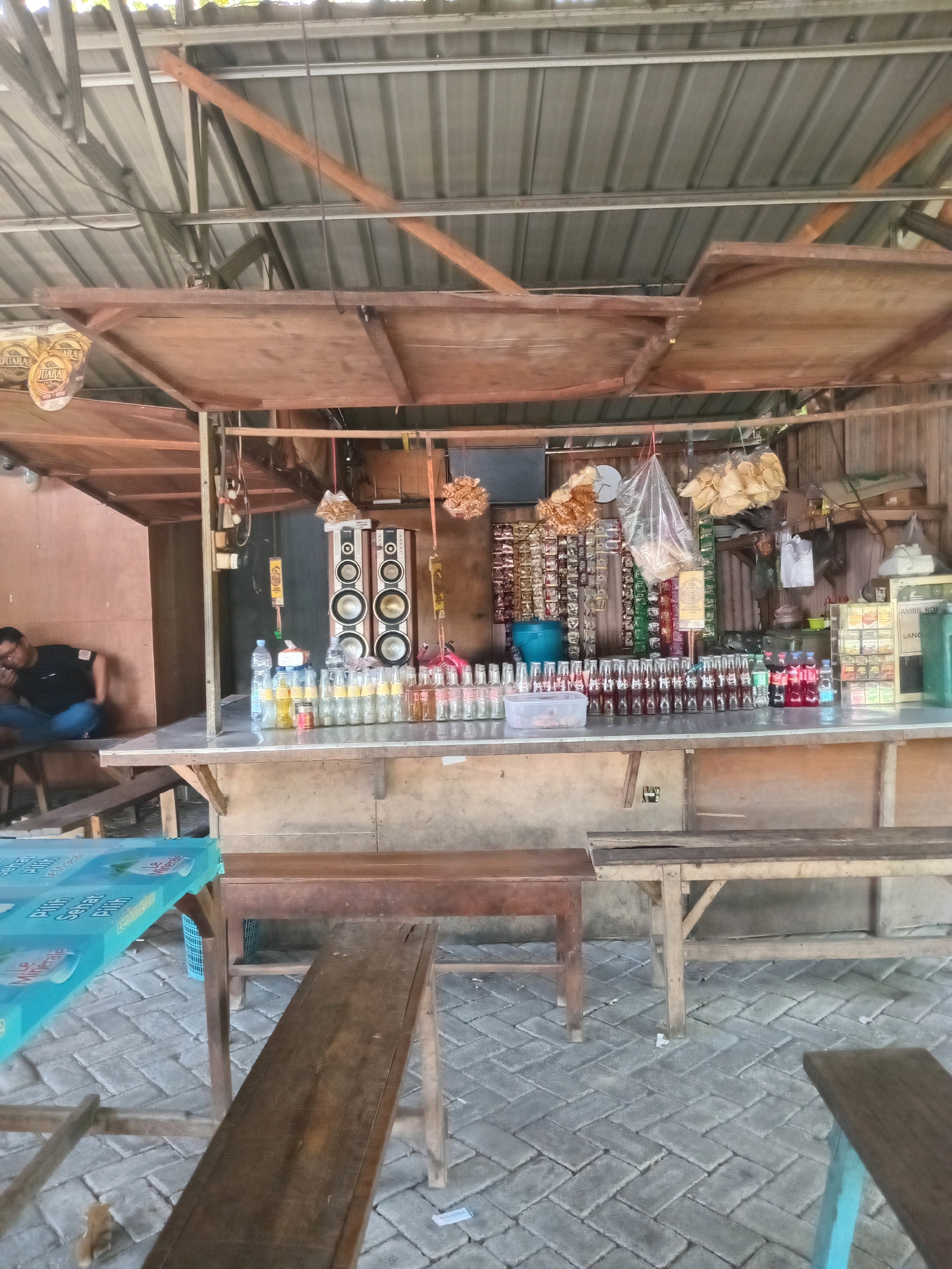 The image depicts an outdoor food stall in Kembangan, Indonesia. The stall has a rustic appearance with a corrugated metal roof supported by wooden beams. The counter is laden with various bottled drinks, including what appear to be soft drinks and water bottles. Behind the counter, shelves are stocked with packaged snacks and other goods. A pair of large speakers is mounted on the back wall. A man in a black t-shirt and blue jeans is seated on a bench to the left of the stall, appearing to be on his phone. The ground is paved with interlocking brick pavers. Several wooden benches and tables are set up in front of the stall for customers. The overall environment suggests a casual, open-air dining or resting area.