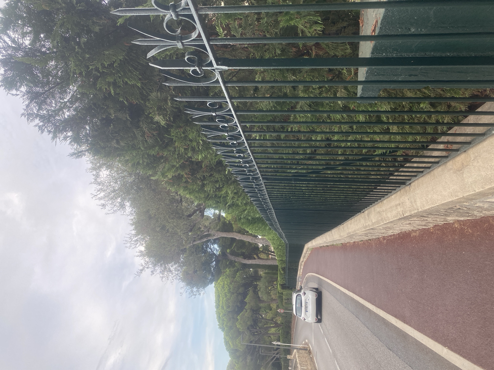 The image shows a road curving to the left in Antibes, France. A white electric car is driving on the road. To the right of the road is a red pedestrian walkway bordered by a low stone wall and a dark green metal fence. Behind the fence, there are numerous cypress trees and other lush green vegetation. The sky is overcast. The scene appears to be a residential area with a scenic road.