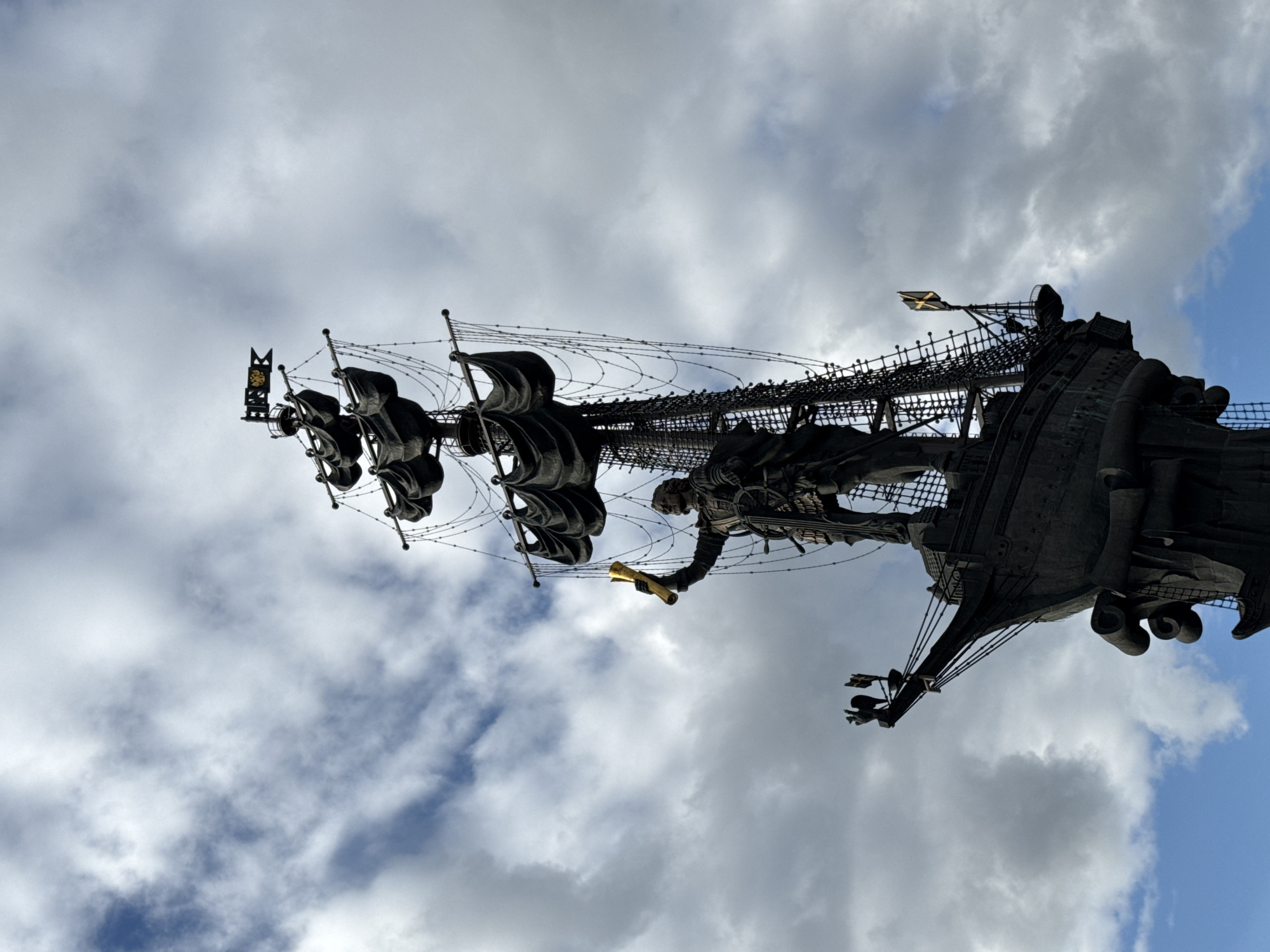 The image shows a low-angle, full-shot view of the Monument to Peter the Great in Moscow, Russia. The monument depicts Peter the Great standing on the deck of a ship, holding a scroll in his right hand. The statue is made of dark metal and is adorned with intricate details, including clothing, armor, and rigging. The ship's masts and sails are also rendered in dark metal, with fabric-like elements suggesting sails. The monument is set against a backdrop of a cloudy sky with patches of blue visible. The overall impression is one of grandeur and historical significance.