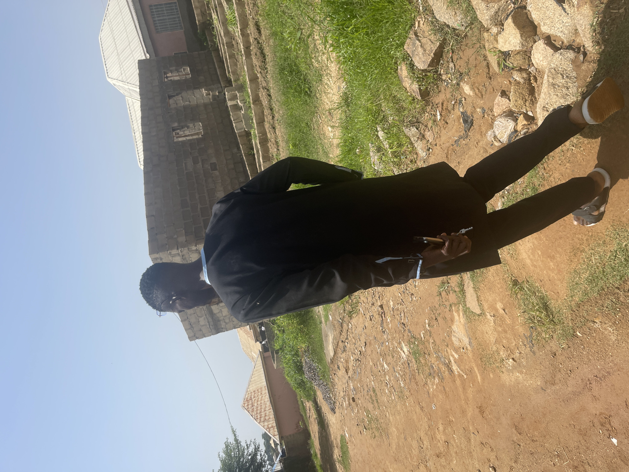 The image shows a person dressed in a black suit and pants, standing outdoors. The person is viewed from behind, with their head turned to the side, looking down. They are wearing glasses and have short, dark hair. The person is holding a small, cylindrical object in their right hand. They are wearing sandals on their feet.
The setting appears to be a somewhat neglected outdoor area. In the background, there is a building constructed from grey bricks with visible windows. To the left of the building, another structure with a corrugated metal roof is visible. The ground is a mixture of dirt, stones, and sparse green grass. There are large rocks scattered around the immediate vicinity.
The sky is clear and blue, suggesting it is daytime, likely sunny. The lighting indicates the sun is relatively high, casting shadows. The overall impression is that of a warm, possibly hot, day. The location context provided is Rafin Par, Nigeria. No text is visible in the image. The scene is static, with no clear indication of a specific event occurring beyond the individual standing and holding an object.