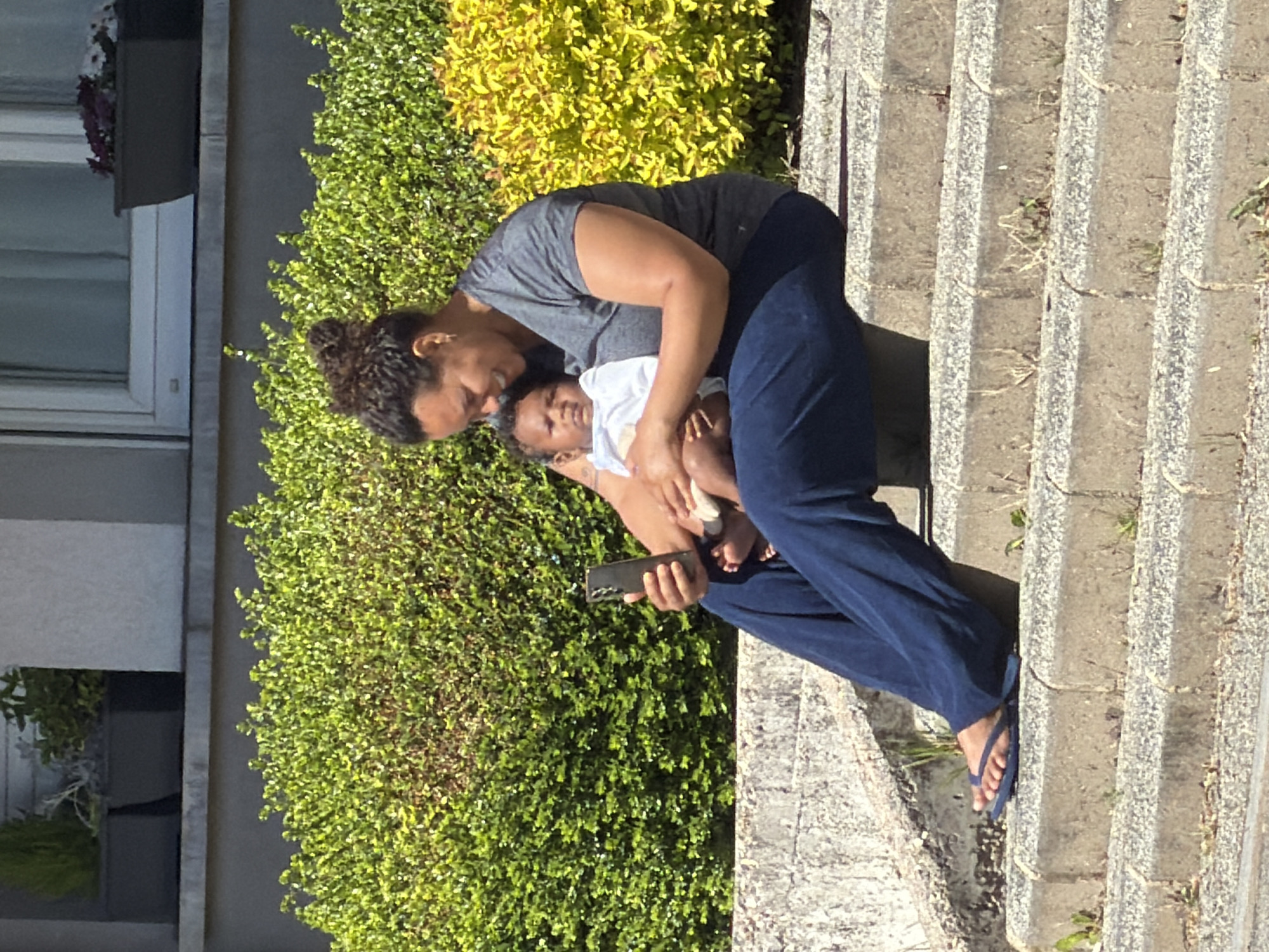 A medium shot depicts a person holding a baby while sitting on concrete steps. The person, who appears to be an adult female, is wearing a gray shirt, blue pants, and blue flip-flops. The baby is dressed in a white top. The woman is holding a black smartphone in her right hand. Behind them is lush greenery, including a yellowish-green bush and a darker green hedge. A building with a window is visible on the left side. The scene appears to be in a residential area in Esch-sur-Alzette, Luxembourg.