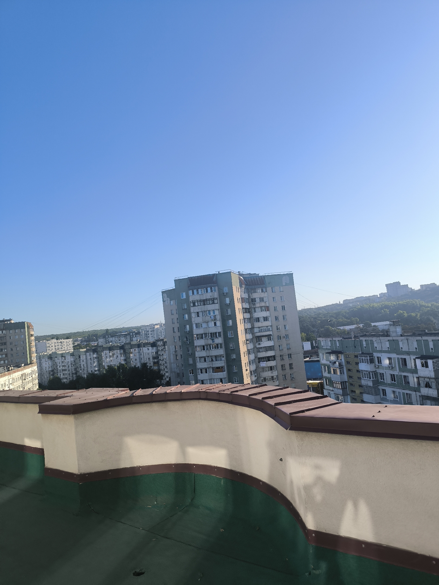 The image is a view from a rooftop in the city of Chișinău, Moldova, looking out over a residential area. The foreground shows a curved rooftop edge with a cream-colored textured wall and a dark green surface below. Beyond the rooftop, a cluster of Soviet-era apartment buildings with light-colored facades and numerous windows dominates the mid-ground. The buildings are densely packed, with some appearing taller and more modern than others. In the background, a lush green hillside rises, dotted with more buildings and trees, with a clear blue sky overhead. The image captures a typical urban landscape of Chișinău, characterized by its communist-era architecture and its proximity to natural green spaces. There are no people or visible activity in the scene.