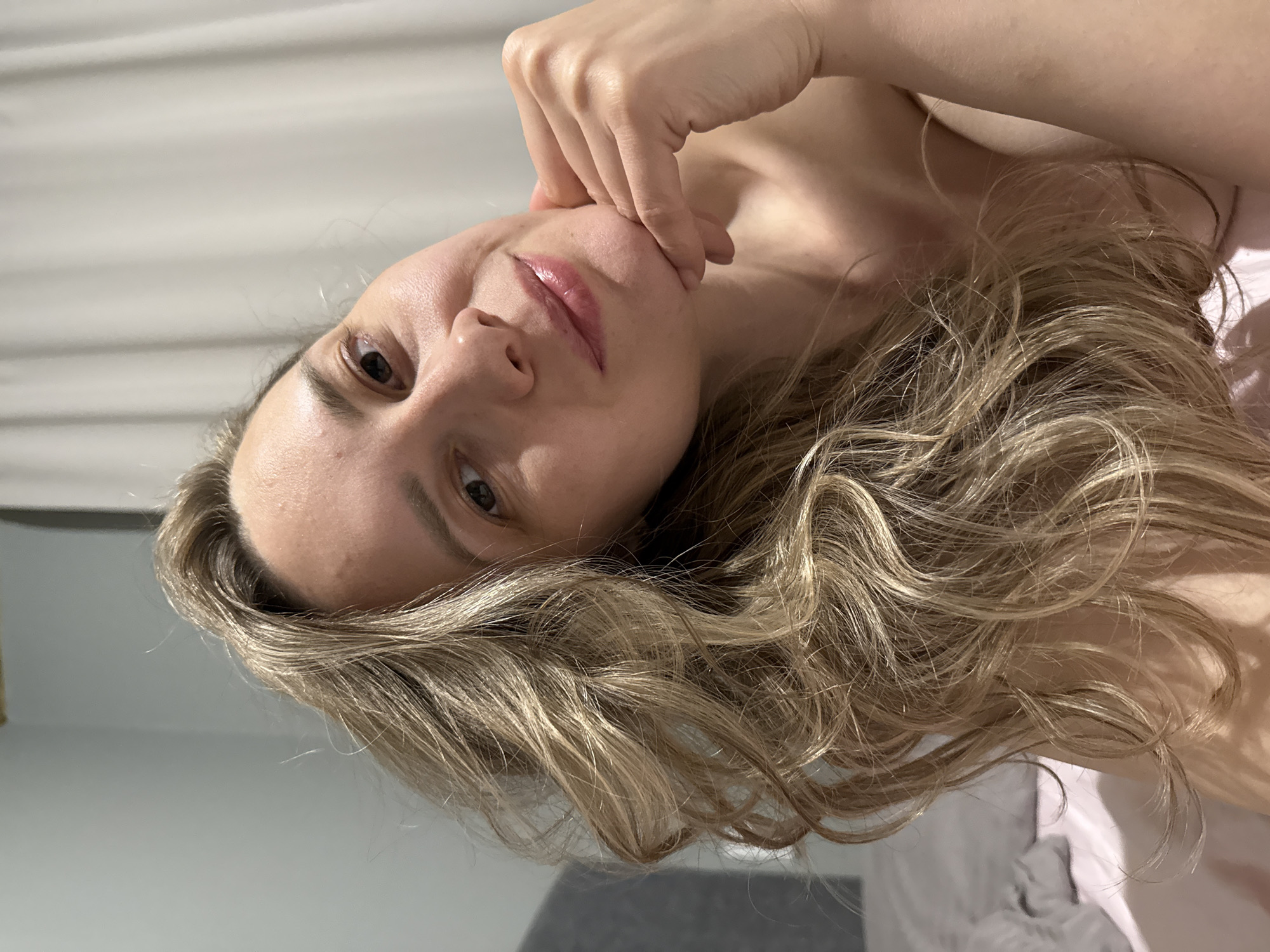 A fair-skinned woman with light brown hair is pictured in an indoor setting. She is propped up, likely in bed, with her hand gently touching her chin. The background shows a light wall and parts of bedding and a gray object, possibly a sofa or another part of the bed. The image provides no indication of the city of Chaikovsky, Russia.
