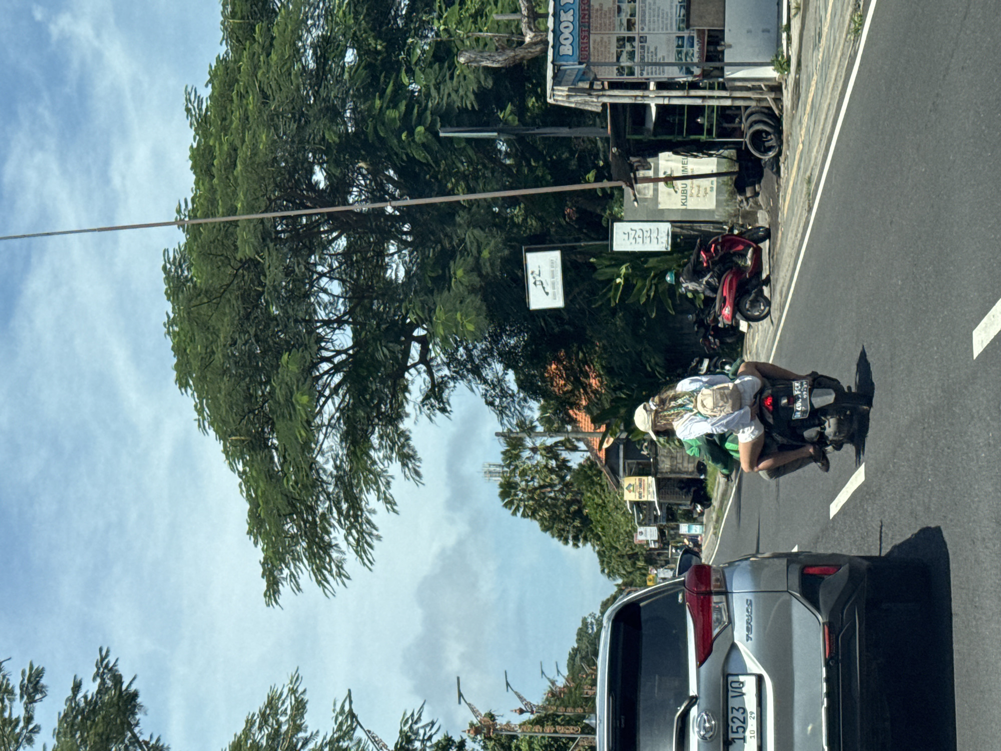 The image shows a street scene in Nusa Dua, Indonesia. A silver Toyota vehicle with the license plate "1523 VQ" is stopped on the asphalt road, and a motorcycle with a rider wearing a white hat and green clothing is passing the vehicle on the right. The motorcycle has a black license plate with white lettering. On the side of the road, there are trees, street signs, and a parked red motorcycle. Signage includes a "BOOK TOURIST INFO" sign and another with "KUBU IMEL" listed. The sky is visible through the trees and is partly cloudy.