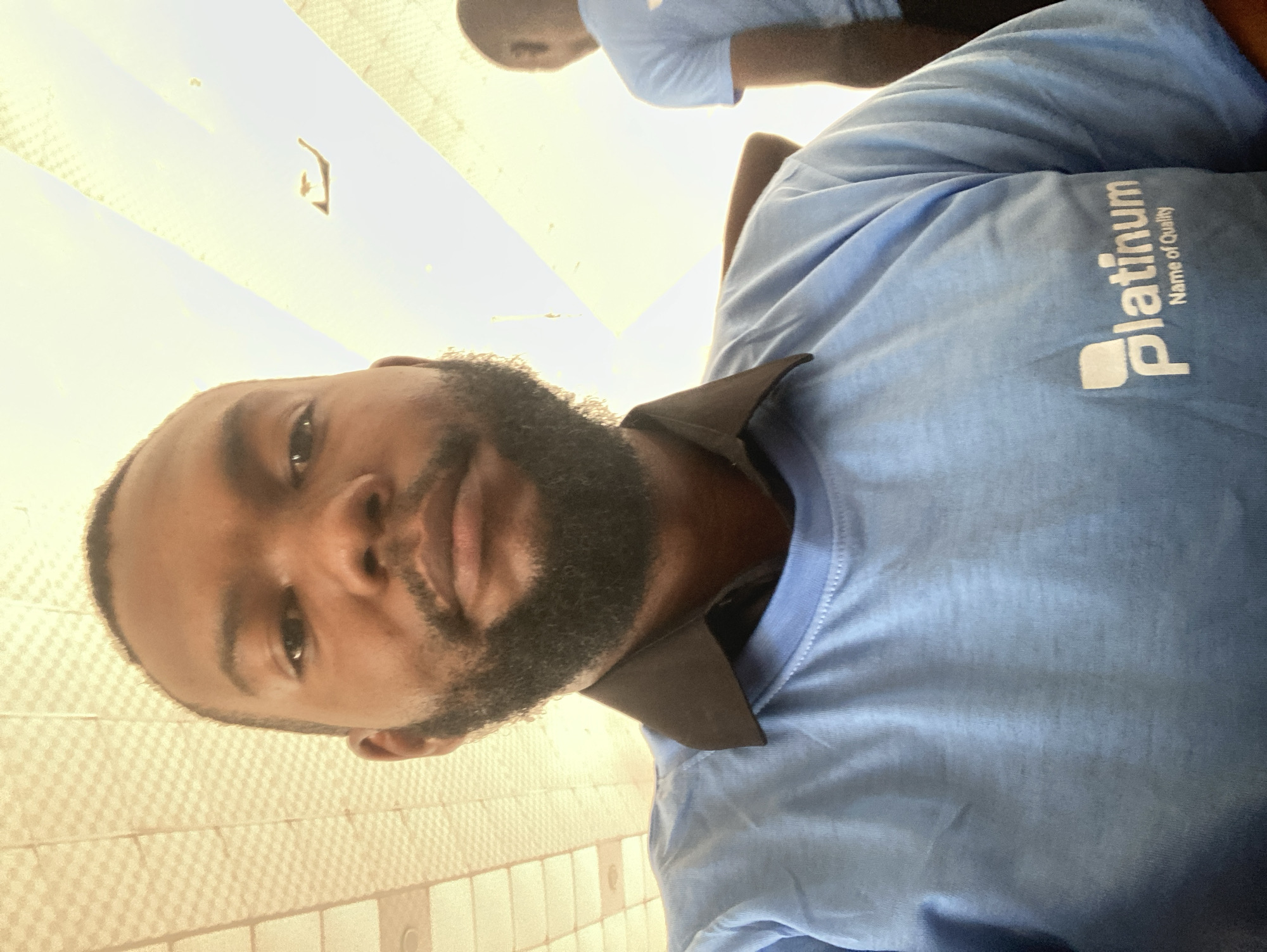 A man with dark skin and a full beard is facing the camera. He is wearing a light blue T-shirt with the word "Platinum" printed on it. The background appears to be a tiled wall. The location is possibly in the city of Jos, Nigeria. There is a blurry reflection of another person and a light source visible in the upper part of the image.