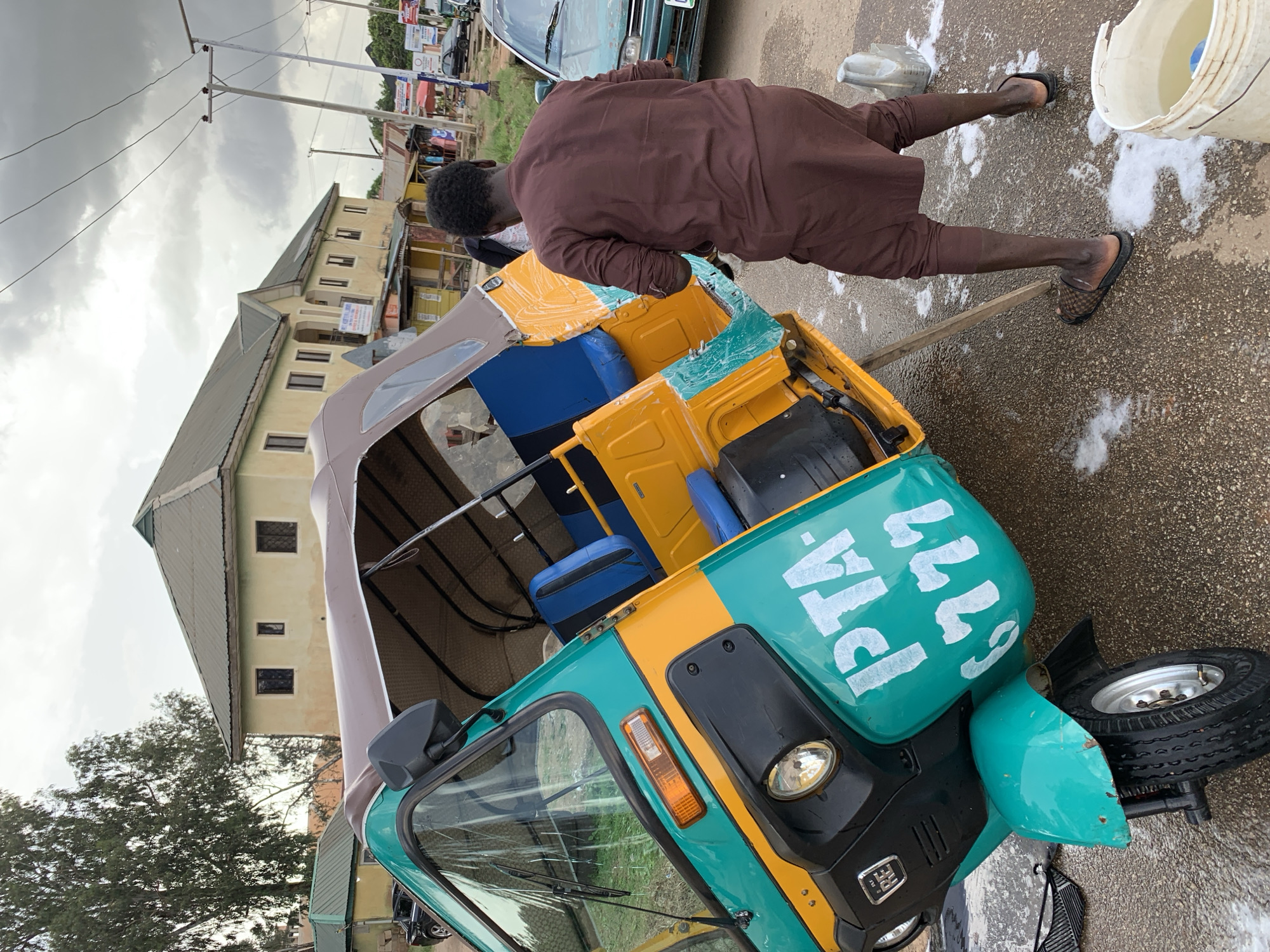 In Jos, Nigeria, a man is cleaning a teal and yellow auto-rickshaw. The auto-rickshaw is tilted on its side, supported by a wooden plank. The man, wearing a brown traditional outfit, is washing the vehicle with a cloth. The ground is wet and soapy, indicating the cleaning process is underway. In the background, buildings and trees are visible, suggesting an urban or semi-urban environment. A green car is parked nearby. The sky is overcast with clouds. The auto-rickshaw has "PTA 6777" written in white on its side.