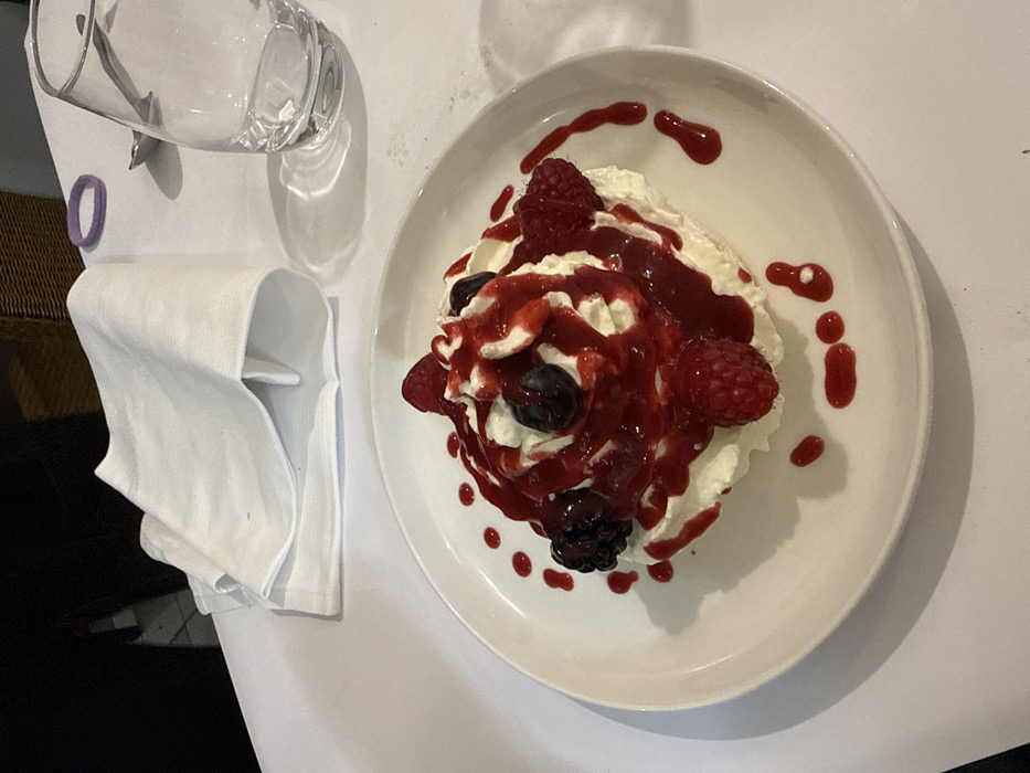 A white plate holds a dessert of whipped cream topped with raspberries, blueberries, and blackberries, drizzled with a red berry sauce. A folded white napkin rests to the left of the plate, next to a glass. The scene appears to be set on a white tablecloth.