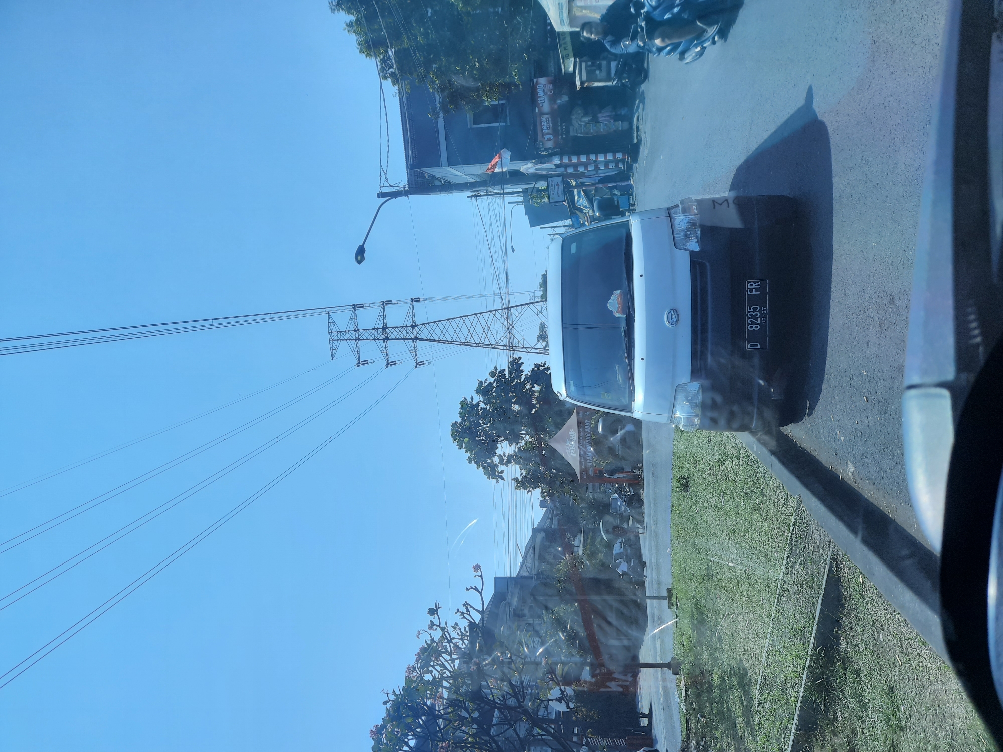 The image captures a street scene in Bandung City, Indonesia, under a clear blue sky. In the foreground, the hood and windshield of a vehicle are visible, suggesting the photo was taken from inside a car. A white van with the license plate "D 8235 FR" is the primary subject, positioned in the middle of the road. The van appears to be stationary or moving slowly.
To the left of the van, a patch of green grass is visible next to a gray curb. Beyond the grass, there are buildings and trees. One building has a prominent tent-like structure with text that is partially obscured but appears to be advertising food. Several people are seen walking or standing in this area, some holding umbrellas.
In the background, a large electricity pylon and high-voltage power lines dominate the skyline, indicating the presence of significant electrical infrastructure. A street lamp is also visible. To the right of the van, more buildings line the street, some with visible signage. A motorcycle with a rider is moving away from the viewer on the right side of the road. The overall impression is a typical urban street scene with traffic and pedestrian activity.