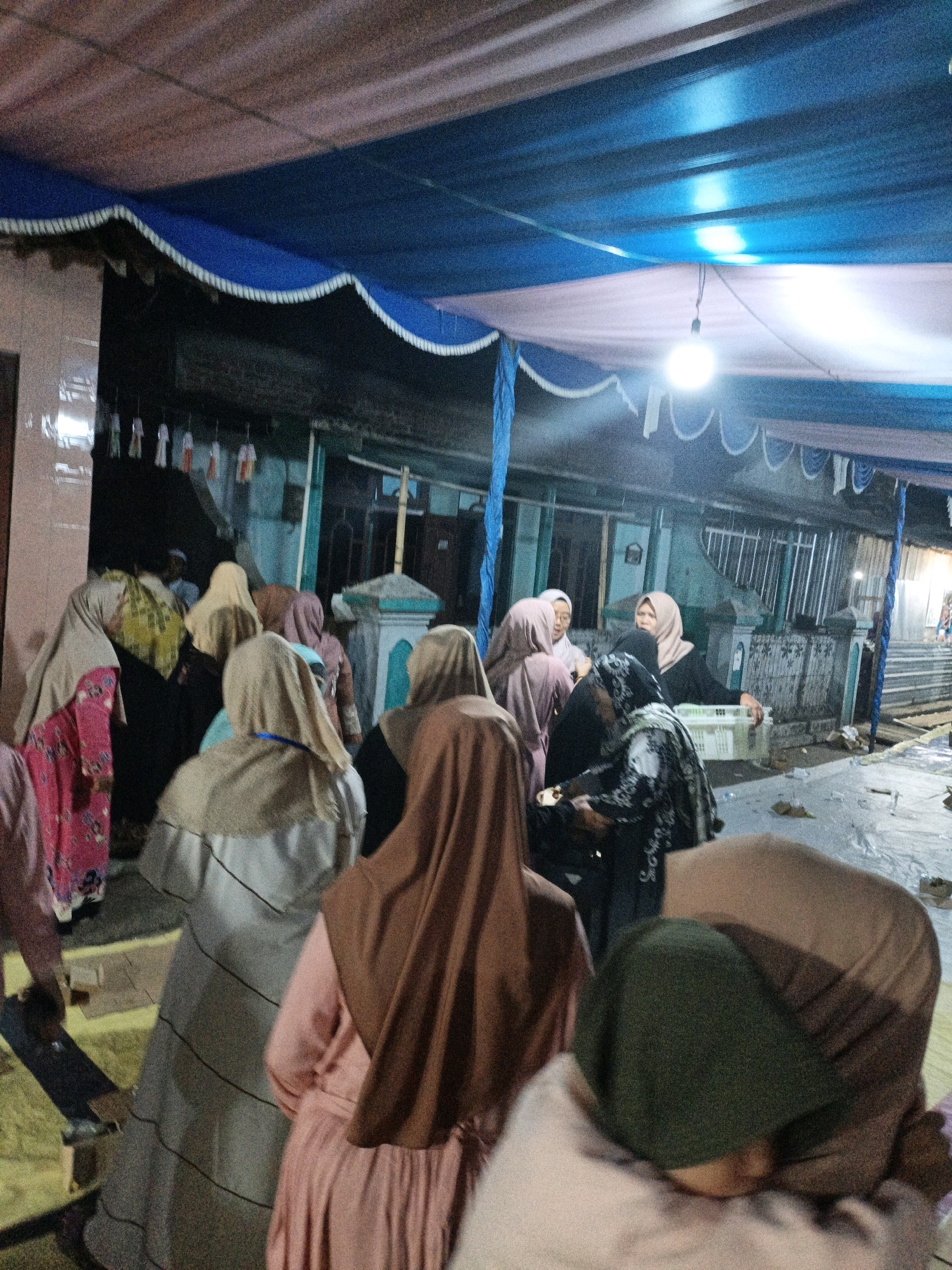 In the city of Tanahbaya, Indonesia, an outdoor gathering is taking place under a decorative canopy. The canopy is constructed from fabric draped from a blue and pink structure, with white trim along its edges. A single light bulb hangs from the canopy, providing illumination. A group of women, most wearing hijabs, are gathered together. Some are standing, while others appear to be in prayer or contemplation. In the background, there are buildings and a fence. The ground is covered with a yellow mat. The overall scene suggests a communal event or religious observance.