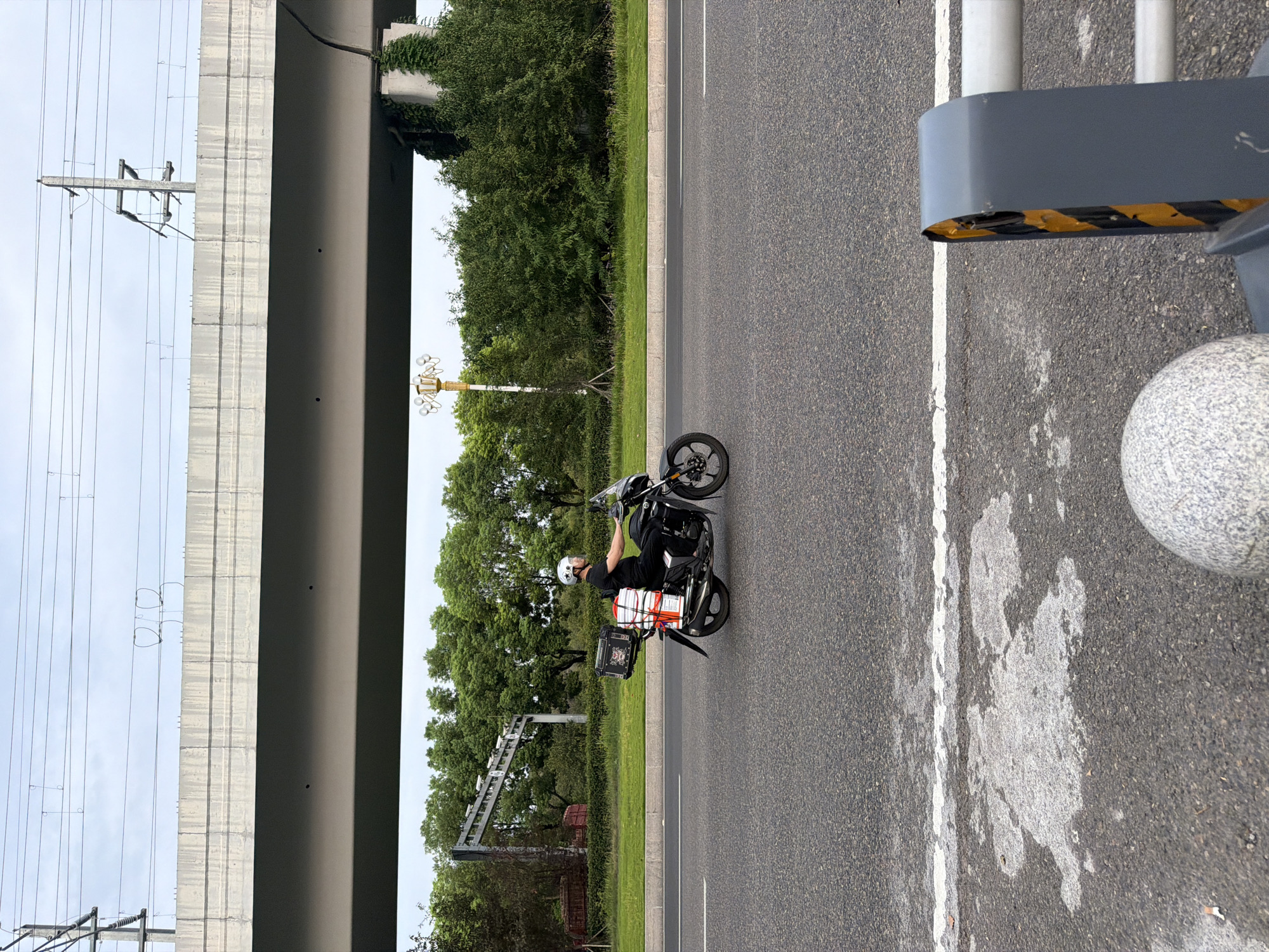 In Jiangjin, China, a man wearing a helmet rides a black motorcycle down a paved road. He is dressed in a black t-shirt and pants. The motorcycle is equipped with a black storage box on the back, containing several white boxes. The road is lined with grass and trees, and an overpass supported by concrete pillars runs across the top of the frame. Overhead, power lines stretch across the sky. In the foreground, a concrete sphere and a traffic barrier are visible. The sky is overcast.