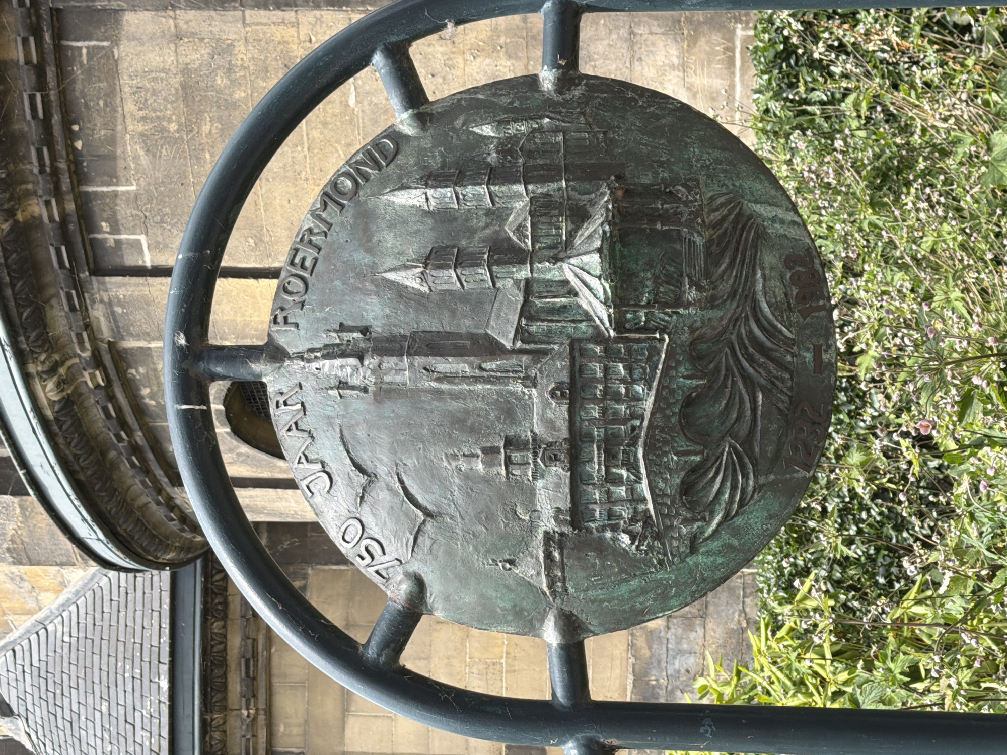 The image shows a circular bronze relief mounted on a metal frame, possibly a historical marker or artwork. The relief depicts a cityscape with buildings and spires, likely representing Roermond. Inscribed around the circumference of the relief are the words "750 JAAR ROERMOND" and the years "1232" and "1982". The relief has a green patina, indicating its age and exposure to the elements. Behind the relief, a stone building with a tiled roof is visible, suggesting an urban or historical setting. In the foreground, lush green foliage with small pink flowers partially obscures the lower portion of the relief. The scene is captured outdoors, with natural light illuminating the artwork and its surroundings. The overall impression is one of historical commemoration and artistic representation within a city.