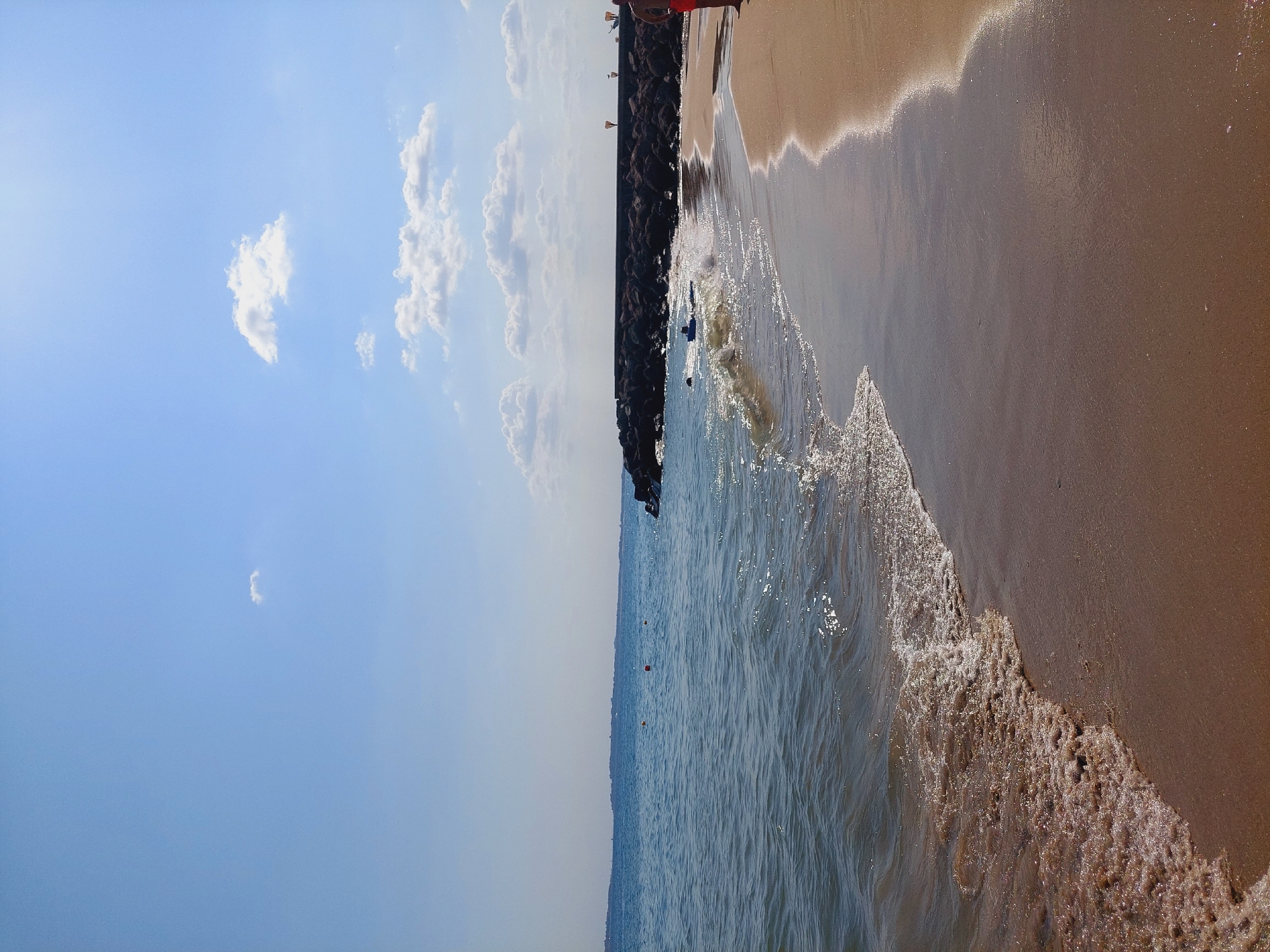 The image depicts a sunny day at a beach in Sveti Vlas, Bulgaria. The foreground is dominated by a sandy beach with gentle waves lapping at the shore. The water is a clear blue, reflecting the bright sky above. In the mid-ground, a breakwater made of large rocks extends into the sea, with a few buoys visible in the water near the shore. Further out, the coastline is visible on the horizon, with a hazy outline of buildings. The sky is a vivid blue, dotted with a few fluffy white clouds. On the right side of the image, a person wearing red shorts stands on the beach, facing away from the camera. The overall scene is tranquil and evokes a sense of a pleasant day at the seaside.