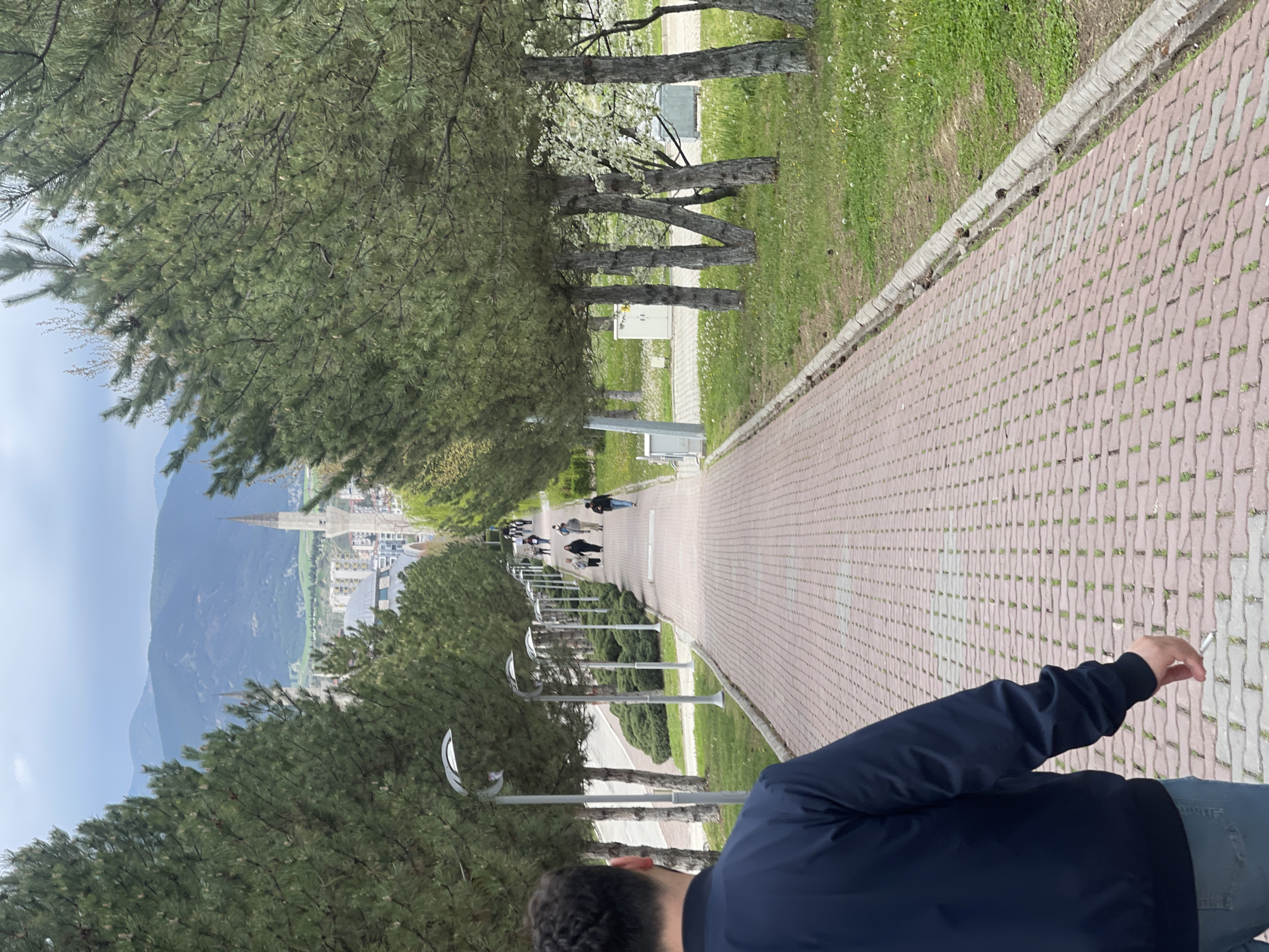 The image, oriented correctly, depicts an outdoor scene in Karabük, Turkey, featuring a paved pathway leading downhill towards a distant town nestled against mountains.
In the foreground, a person is seen from behind, wearing a dark blue jacket and light blue jeans. Their right hand, holding a lit cigarette, is visible on the right side of the frame. The person has dark, curly hair.
The pathway is constructed from red, brick-patterned interlocking pavers, with green grass and scattered white flowers bordering it. A concrete curb separates the path from the grassy areas. The path is lined on both sides by numerous evergreen trees, likely pines, with thick canopies providing shade. Modern-looking street lamps with curved, silver-colored tops are regularly spaced along the path.
Further down the path, several groups of people are seen walking, appearing as smaller figures in the distance. On the left side of the path, nestled among the trees, are low, light-colored utility boxes.
In the midground and background, the path descends into a valley where a town or city is visible. Prominent among the buildings is a tall, slender structure with a pointed spire, resembling a minaret. Beyond the town, layers of hazy, blue-tinged mountains stretch across the horizon.
The activity taking place is people walking along the path, with the foreground individual also smoking. The scene appears to be set during daytime, under a partly cloudy or hazy sky, suggesting mild weather. There is no visible text in the image.