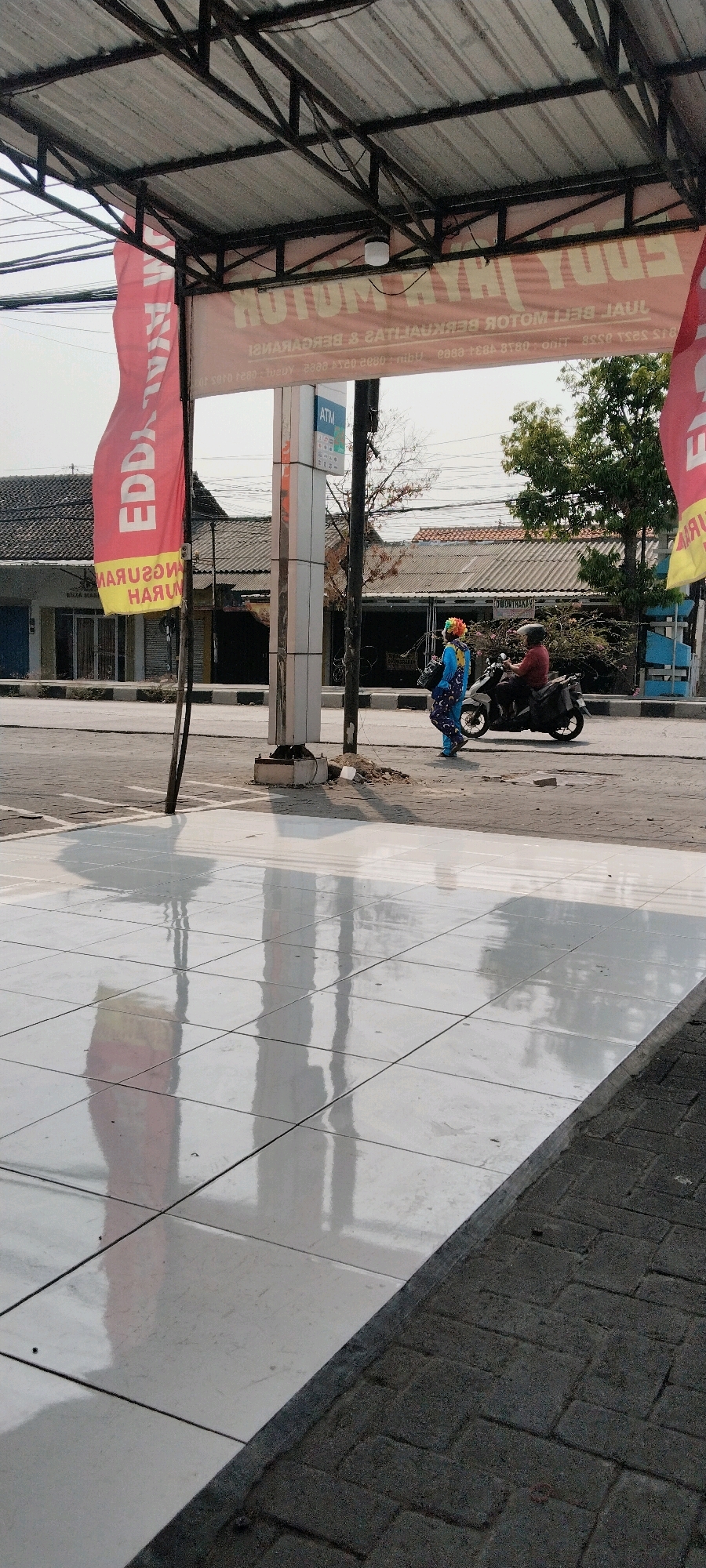 The image depicts an outdoor scene in Semarang, Indonesia, possibly near a street or marketplace. In the foreground, a polished white tiled surface reflects the surroundings, including overhead structures and the sky. A man wearing a maroon shirt and a dark helmet rides a black motorcycle, moving away from the camera on a paved road. Beside the road, to the left of the motorcycle, a person in a colorful clown costume, with a blue outfit and a wig of red, yellow, and green hair, walks towards the motorcycle, carrying a dark bag. Behind them, an ATM machine is attached to a tall, light-colored pillar. A large red banner with Indonesian text, partially obscured by the pillar, hangs vertically. Another banner, predominantly orange with dark text, is stretched horizontally above, also displaying Indonesian writing. The background shows buildings with tiled roofs and some trees, suggesting an urban or semi-urban setting. The overall lighting suggests it is daytime, with shadows visible on the tiled surface.