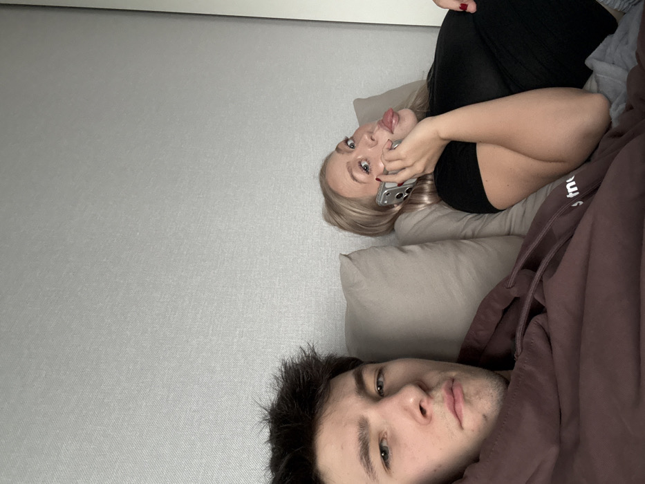 In a residential setting within Yanino-1, Russia, two individuals are depicted reclining indoors. On the left, a young adult male with dark, disheveled hair and a neutral expression looks directly at the camera. He is wearing a dark maroon or brown hooded sweatshirt. To his right, a young adult female with blonde hair is holding a silver-colored smartphone to her right ear with her right hand, her left hand also visible with red nail polish. She is wearing a black top and is captured mid-expression, with her tongue slightly extended. Both individuals are positioned against light-colored pillows and a textured light grey wall. A white vertical architectural element is visible on the far right edge of the frame.