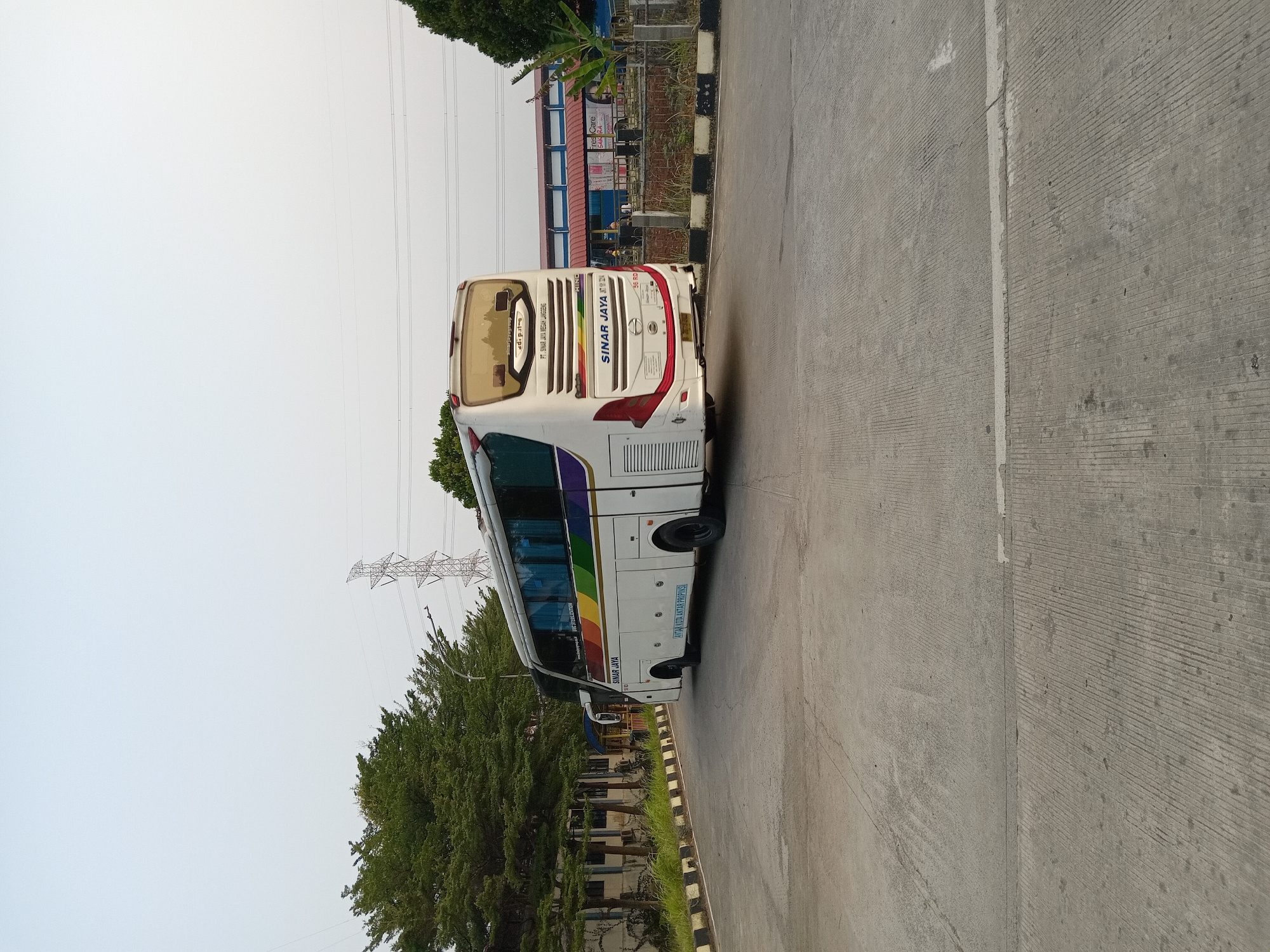 A wide shot captures a white intercity bus with rainbow-colored stripes on its side parked on a paved road in Depok, Indonesia. The bus is branded with "SINAR JAYA" and other text, including a license plate that reads "B 7012 BZA." In the background, a building with a red roof and signs, including one for "FREE CARE," is visible. Trees line the left side of the road, and a power line tower stands tall in the distance. The sky is a pale, overcast gray.