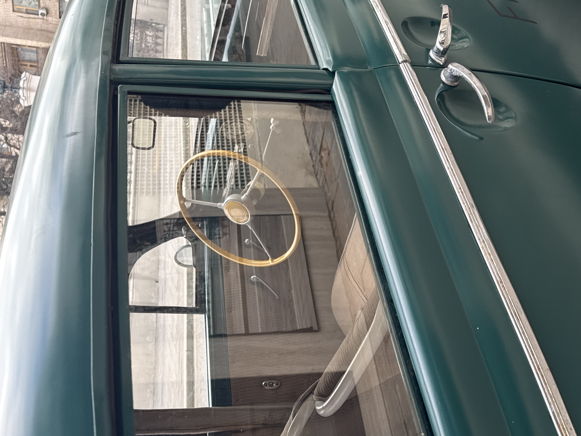 A close-up, angled shot captures the side of a vintage dark green public transport vehicle, likely a bus or trolleybus, in Moscow, Russia. The view is partially obscured by reflections in the vehicle's windows. Through the glass, a classic wooden steering wheel with a metallic center is clearly visible in the foreground of the interior. Further inside, light-colored, possibly striped, seats can be seen. The exterior of the vehicle features two shiny chrome door handles and a horizontal chrome trim strip running along the side. Reflections in the windows show glimpses of an urban environment, including architectural details of nearby buildings and bare tree branches, indicating a daytime setting. The vehicle appears to be well-preserved, suggesting it could be a restored model or part of an exhibit. No specific activity is taking place, and no discernible text is visible on the vehicle.