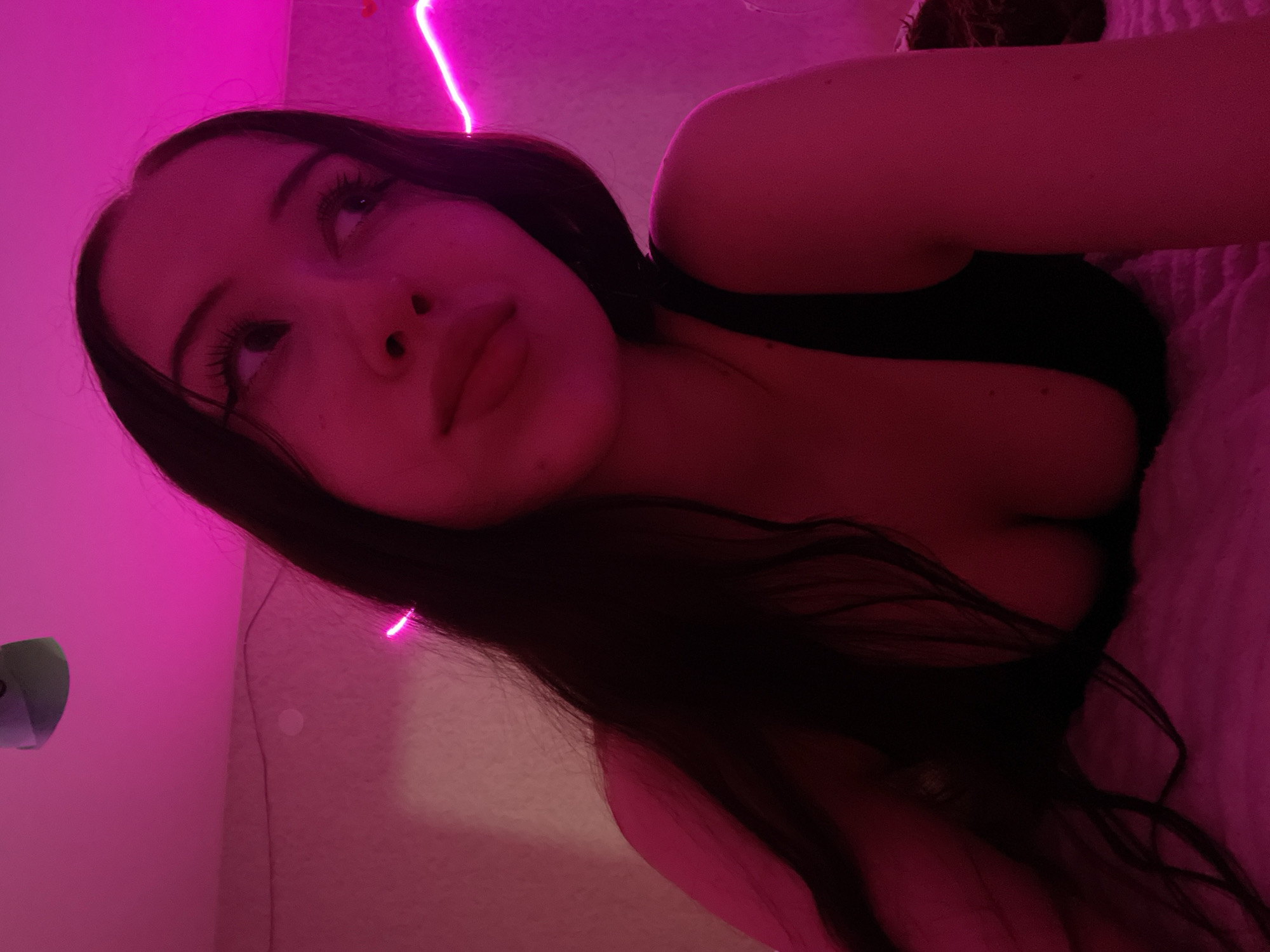The image is a close-up, angled shot of a young woman in dim lighting with a pink neon glow. She has long, dark hair and is wearing a black top with a low neckline. The background is blurred and appears to be a bedroom setting, with a textured surface, possibly a duvet or blanket, visible on the right side. A bright pink neon light strip is visible along the top left of the frame, casting a strong pink hue over the scene and reflecting on the woman's hair and skin. The woman's face is partially visible, with her eyes looking upwards. Her expression appears contemplative or relaxed. The overall mood is intimate and atmospheric, enhanced by the low light and vibrant neon illumination. No text is visible in the image. The location context of Votkinsk, Russia, does not provide any specific visual cues within the image itself.