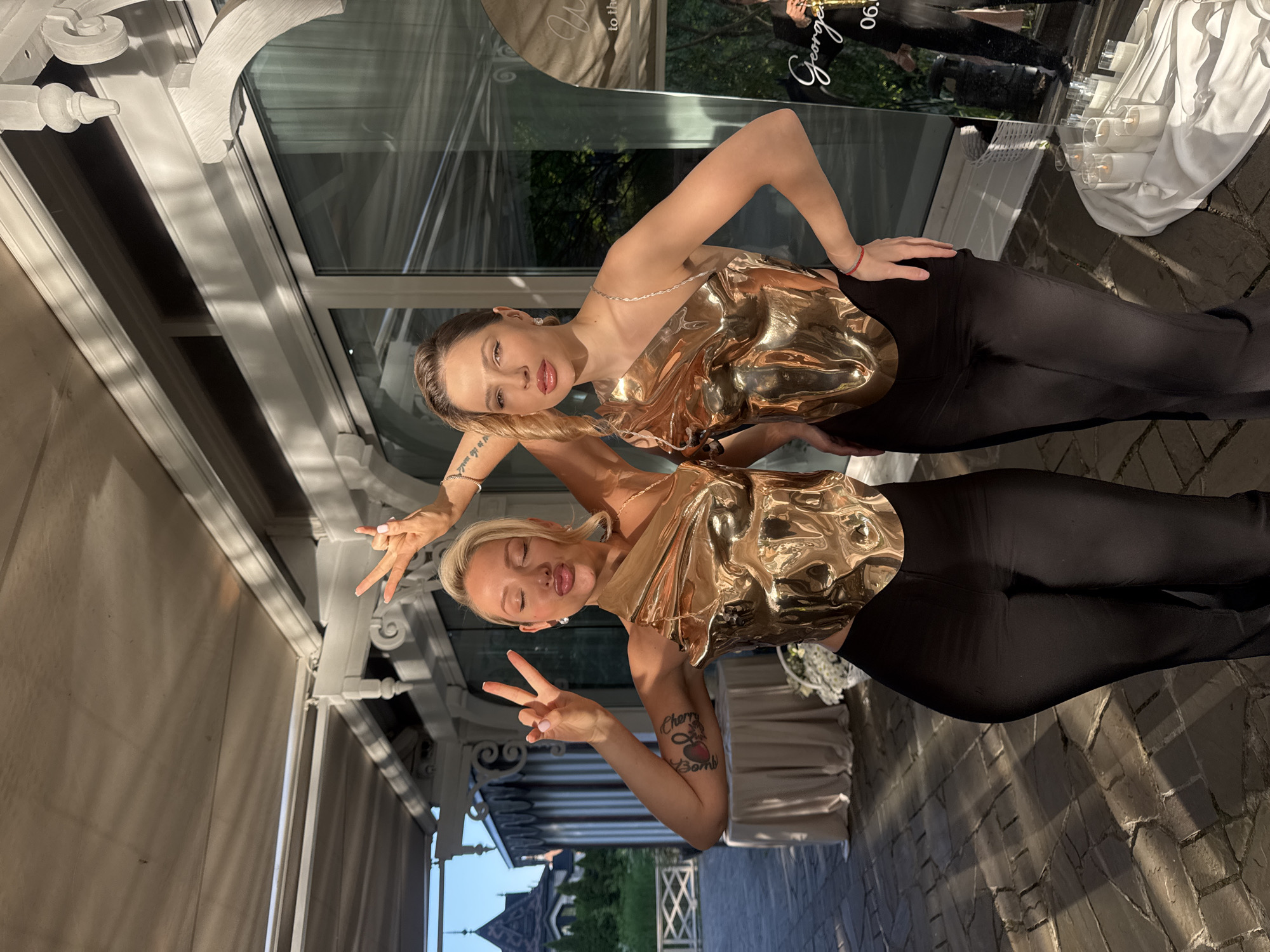 This image depicts two women posing outdoors in Sel'skaya Nov', Russia, during daylight hours, likely late afternoon or early evening due to the long shadows. Both women are wearing reflective gold-toned, form-fitting tops and black trousers.
The woman on the left has blonde hair, styled away from her face. She has her eyes closed and lips pursed in a kissing expression. Her right hand is raised, forming a "peace" sign with her index and middle fingers. Her left arm is bent, with her hand positioned near her head, also forming a "peace" sign. A tattoo, appearing to be a red heart with text, is visible on her left upper arm. She wears an earring in her visible right ear and a bracelet on her left wrist.
The woman on the right has light brown hair, styled back with a wave near her left temple. She looks directly at the camera with an unsmiling, direct expression. Her right hand rests on her hip. Her left arm is bent, with her hand positioned behind her head. Her gold top features a thin chain strap on her left shoulder. She wears an earring in her visible right ear and a red bracelet on her right wrist.
They stand on a paved surface composed of dark, irregularly shaped stones. In the background, a white architectural structure with large glass windows and decorative white columns, arches, and brackets is visible. Through the windows and beyond, green foliage and trees are apparent. To the far left, a building with a dark, multi-gabled roof and spires is partially visible. To the left of the women, a small round table covered with a light-colored cloth is present. To the right of the women, a large reflective surface, possibly a mirror or sign, shows partial text including "George..." and "06.2". In front of this surface, several white pillar candles in glass holders are arranged on a white cloth on the ground, alongside smaller glass containers. A white basket containing white flowers is partially visible on the ground between the women. The scene is lit by natural light, casting distinct shadows on the ground.