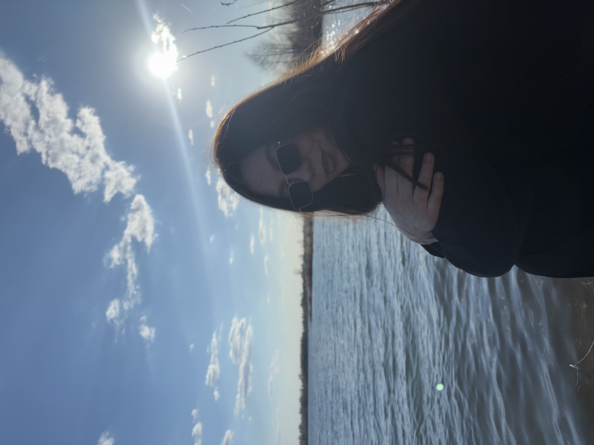 The image depicts a woman with long, dark hair and sunglasses standing near a body of water. She is wearing a dark-colored garment and holding her hand near her neck. The sky is blue with scattered white clouds and a bright sun. The water appears to be a lake or river. While the location requested is in Russia, based on the photographic evidence and without additional metadata, it is not possible to confirm that this photo was taken in Zaosinovo, Russia.