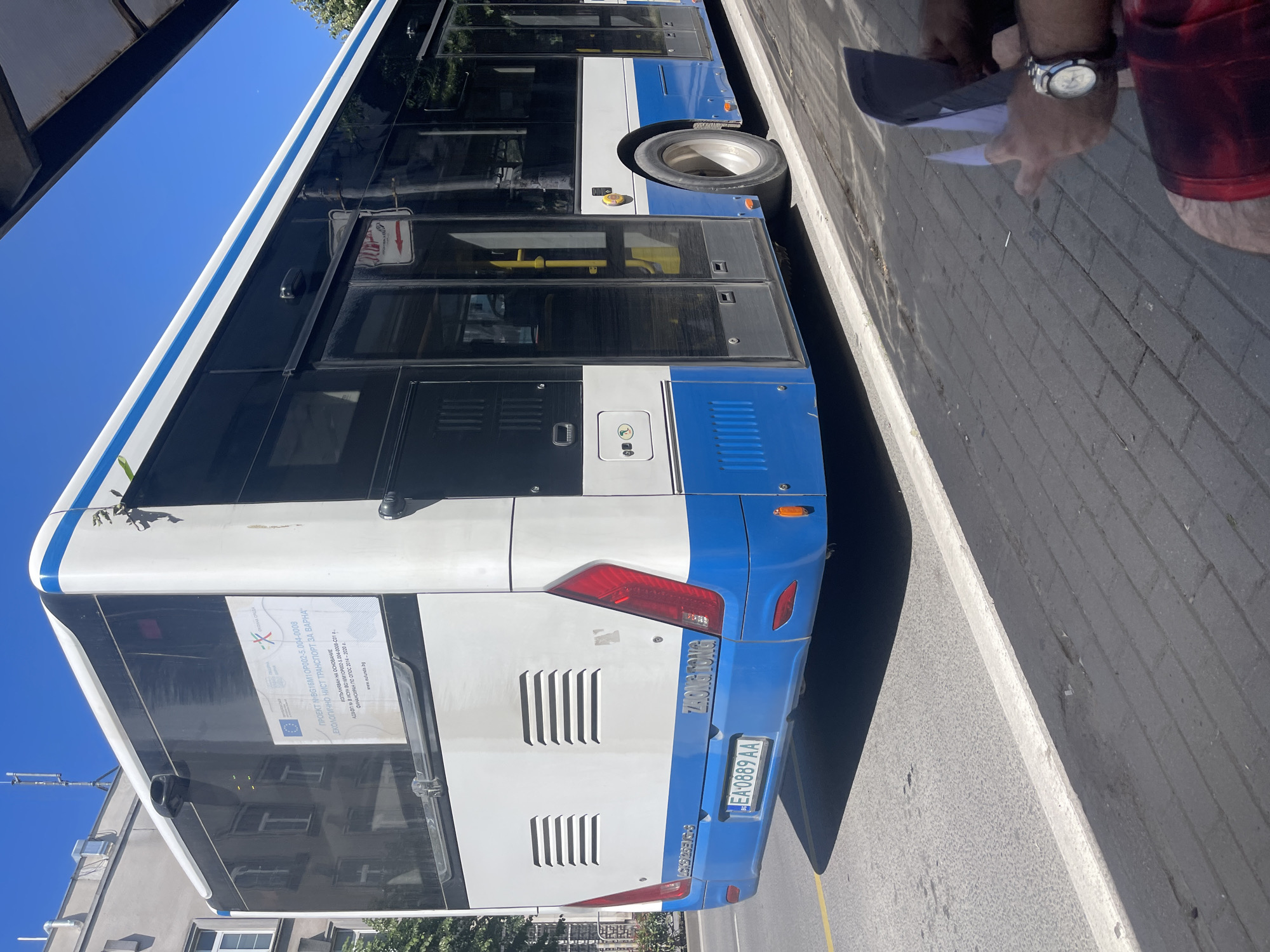 The image shows the rear and side of a blue and white bus, specifically a "Zhong Tong" model, parked on a street in Varna, Bulgaria. The bus has a license plate that reads "EA 0889 AA". A sign on the rear of the bus indicates it is part of a project to improve public transport in Varna, funded by the European Union. In the foreground, a person's legs and hands are visible, holding papers and wearing a watch, suggesting they are waiting at a bus stop. The background shows a building and a clear blue sky.