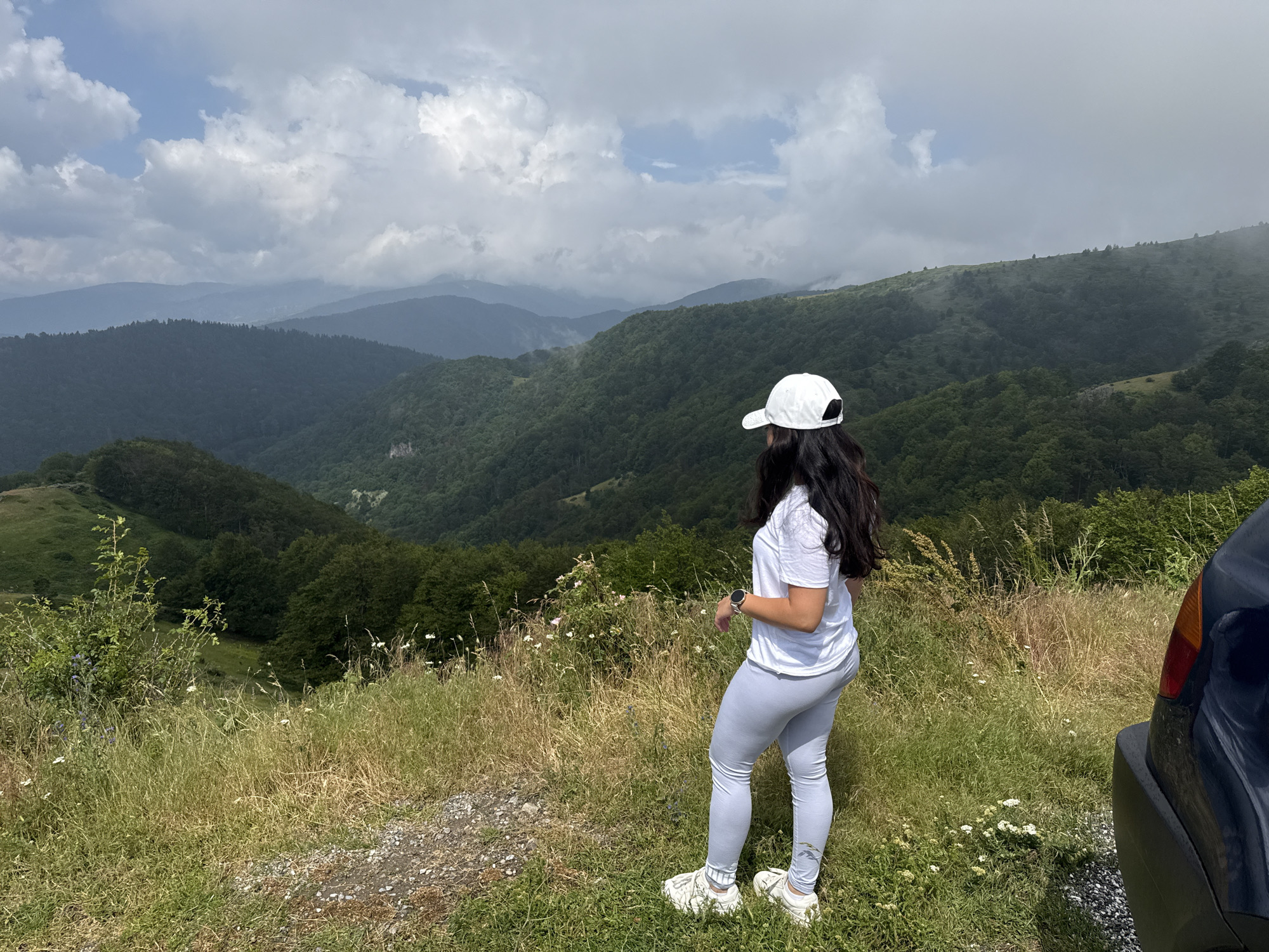 The image shows a person wearing a white cap, white t-shirt, and light gray leggings, standing on a grassy hillside overlooking a valley filled with green trees. The person is facing away from the camera, looking at the landscape. In the foreground, there is dry grass and some white wildflowers. To the right, a dark-colored car is partially visible. The background features rolling hills covered in dense forest, with layers of mountains fading into the distance under a cloudy sky. The location is identified as Hristo Danovo, Bulgaria.
