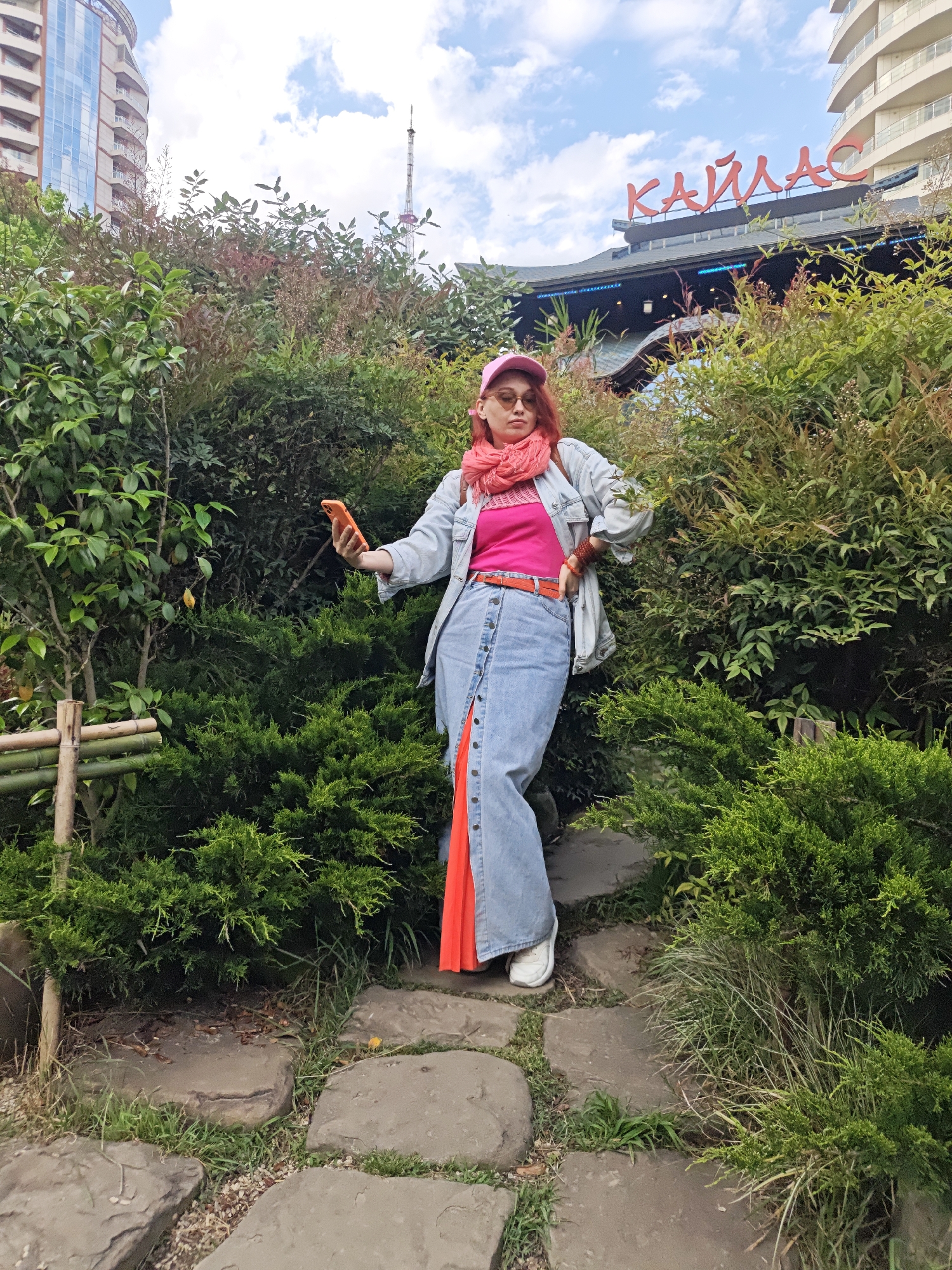A woman stands in a garden, holding a phone in her left hand, appearing to take a selfie. She is wearing a pink hat, a pink top, a long denim skirt with orange accents, a light blue jacket, and white sneakers. An orange scarf is around her neck, and she has an orange belt.
She is positioned on a stone pathway surrounded by dense green bushes. Behind her, a building is visible with the word "KAYLAS" in red letters. Other buildings can be seen in the background as well as a tower.
This photograph was taken in Sochi, Russia.