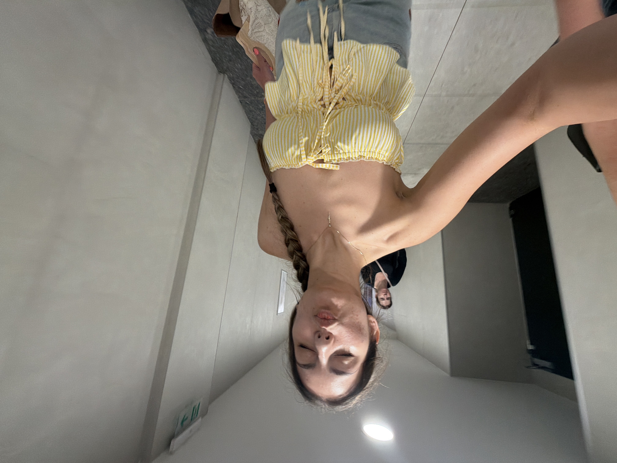 An inverted image shows a young woman taking what appears to be a mirror selfie in an indoor setting. Her head is towards the bottom of the frame, with her body extending upwards. She has long brown hair styled in a braid, closed eyes, and slightly pursed lips. She is wearing a yellow and white striped, off-the-shoulder crop top with drawstring details and light-colored bottoms, possibly jeans or shorts. A small golden necklace with a pendant is visible, along with a belly button piercing. One hand has nails painted a light coral color.
Behind the woman, in a reflection on a mirror or glass surface, is another person, a man with short dark hair and a dark shirt, looking towards the camera's direction. The setting appears to be a bright, modern interior with light grey walls and a light-colored, possibly tiled or polished concrete, floor. A circular white light fixture is visible on the ceiling. Towards the left of the image, near the wall, is a green emergency exit sign with white symbols of a running person and an arrow pointing to the left. The overall impression is of a casual moment captured in a public or semi-public indoor space in Burgas, Bulgaria.