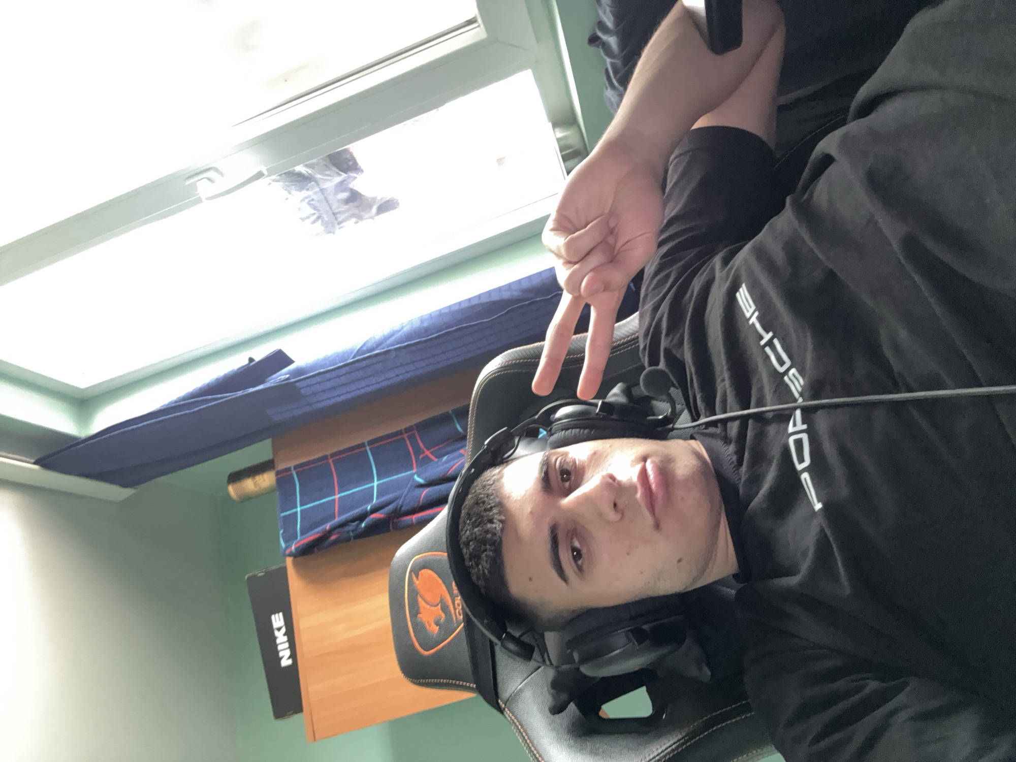 A young man is sitting in a black gaming chair, wearing a black t-shirt with "PORSCHE" printed vertically in white. He has headphones on and is making a peace sign with his right hand. A window is visible in the background, with a partially visible blue and red plaid blanket and a wooden surface below it. A Nike shoebox is visible on the wooden surface. The location appears to be indoors, possibly a bedroom. The city of Sofia, Bulgaria cannot be ascertained from the information provided in the image.