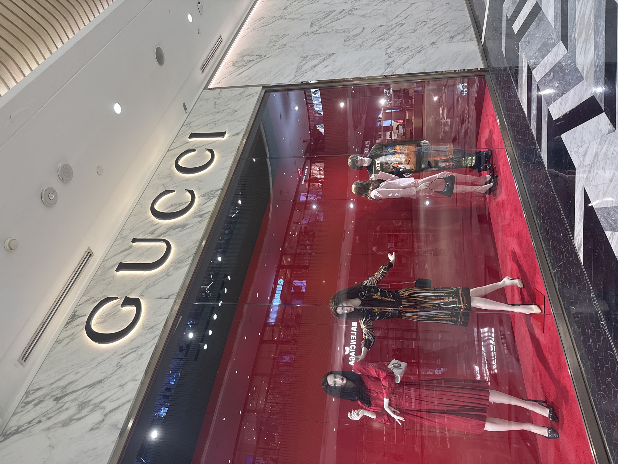 The image shows the exterior of a Gucci store in Wichit, Thailand. The store has a large glass window displaying mannequins dressed in designer clothing. The Gucci logo is prominently displayed above the window. The store is located in a modern shopping mall, with marble flooring and a wooden ceiling. The mannequins are arranged on a red carpet. There are reflections of the mall's interior in the store window. The lighting is bright, and the overall scene is one of luxury and high fashion.