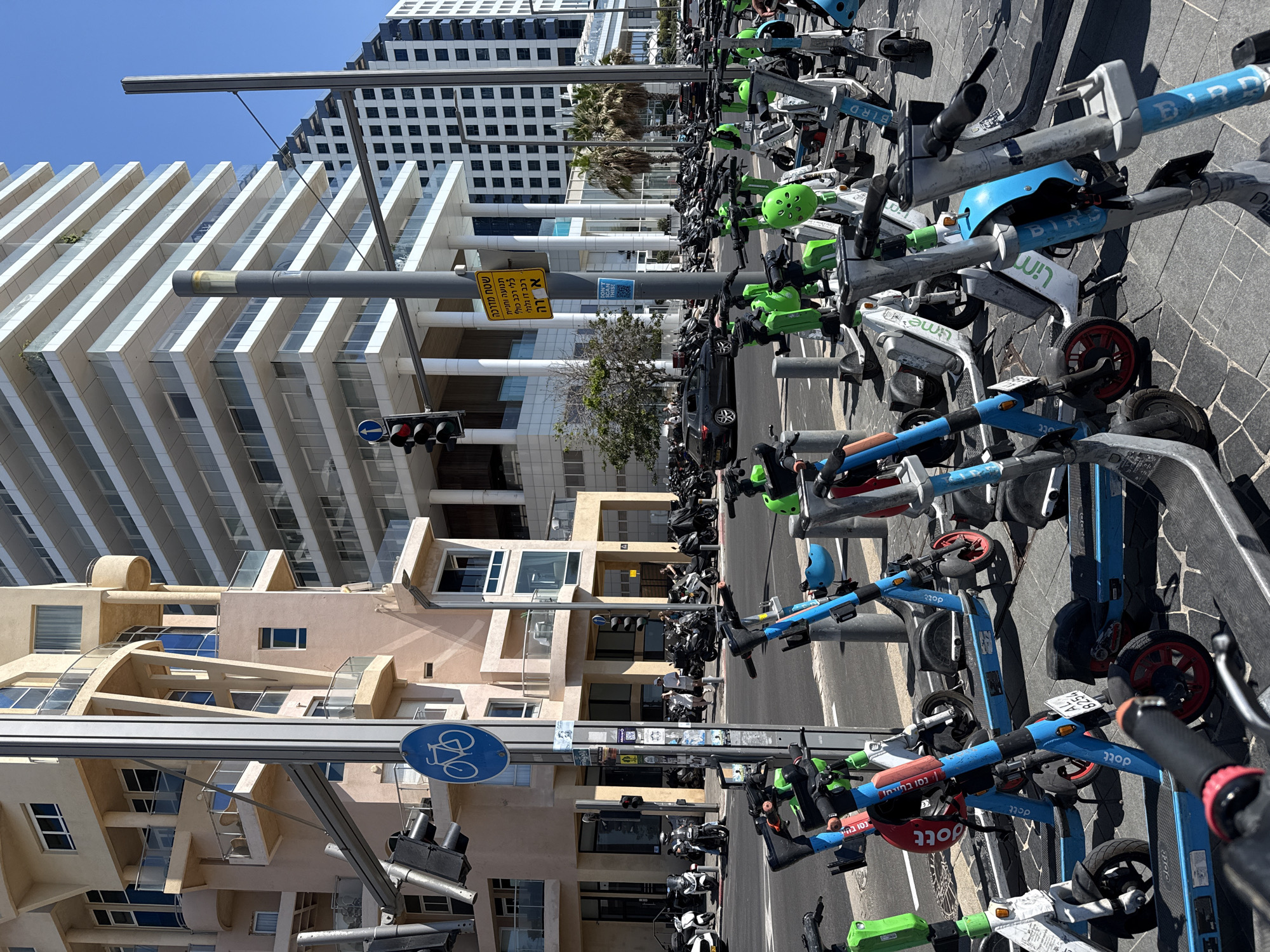 The image captures a street scene in Tel-Aviv, Israel, with a focus on a row of parked electric scooters from various companies like Bird, Lime, and Dott. These scooters are parked alongside a street, seemingly near a designated bike lane, marked by a blue sign. The backdrop features diverse architectural styles, including modern high-rise buildings with geometric designs and structures with arched details. A traffic signal is visible, and street signs in Hebrew and English indicate local regulations. The overall scene depicts urban transportation and architectural diversity in Tel-Aviv.