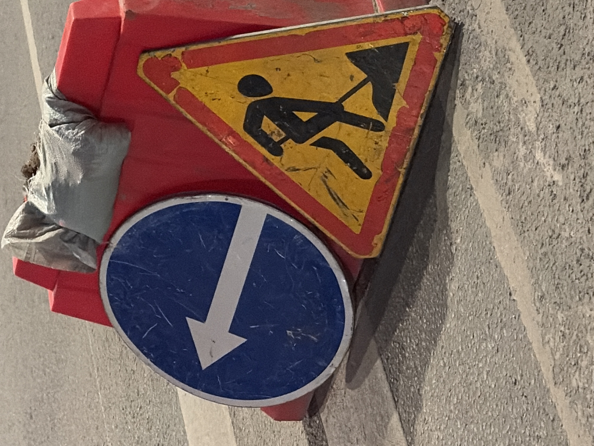 A red traffic barricade sits on asphalt, supporting two signs. The upper sign is triangular and yellow with a red border, displaying a black silhouette of a person digging. Below it is a round blue sign with a white arrow pointing down. To the side of the barricade is a crumpled gray bag. The scene is on a street in Perm, Russia.