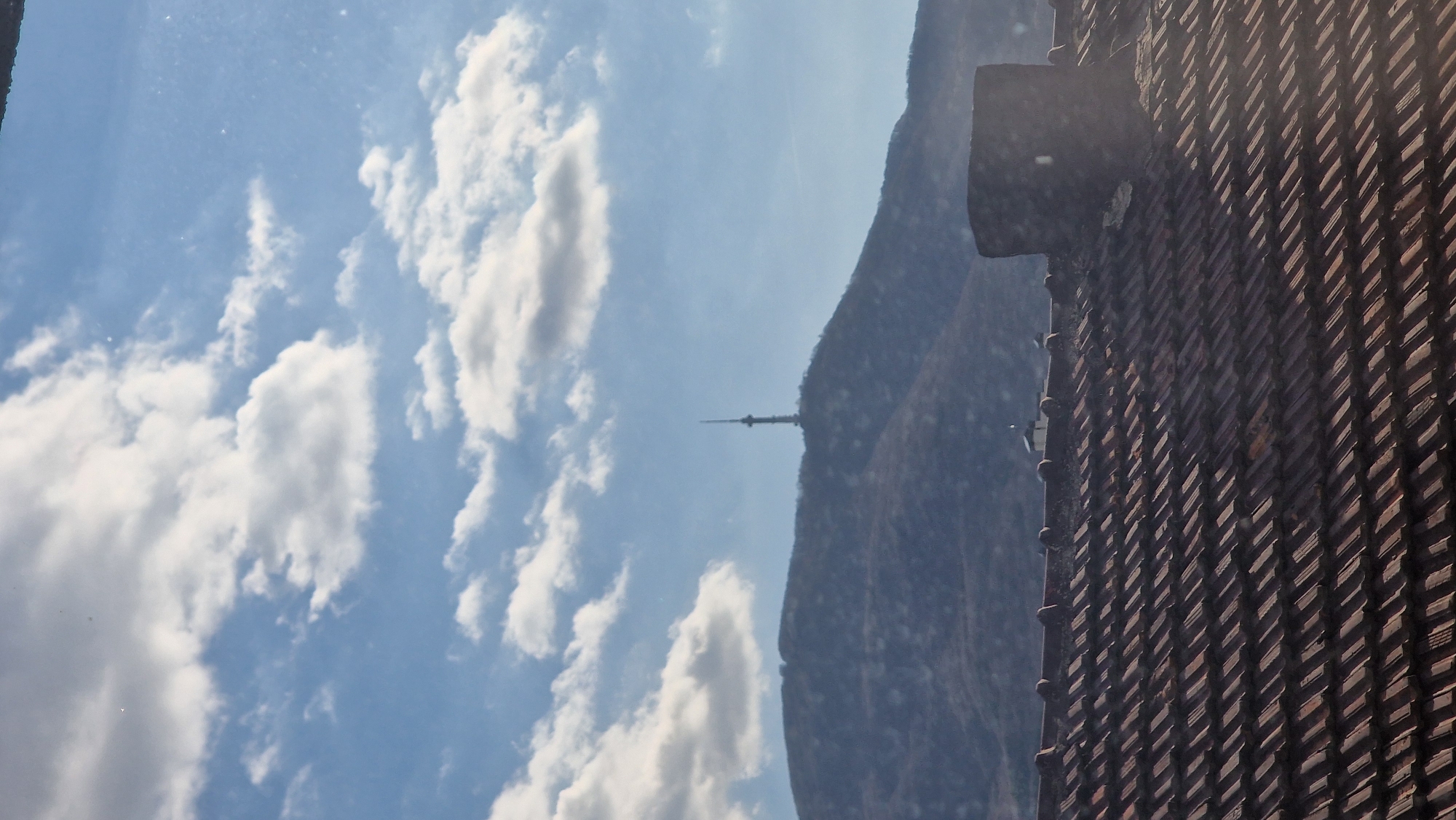 A slightly rotated photograph taken from an elevated position features a view of Sofia, Bulgaria. To the left, a sky with scattered clouds suggests a bright day. In the center, a mountain partially obscured by haze bears a distinctive radio tower on its summit. To the right, the corner of a building with a steeply pitched roof covered in patterned tiles dominates the foreground. The close proximity of the building element suggests the photo was taken from inside a structure, potentially a window or balcony of an adjacent building. The viewpoint offers a vista of the surrounding landscape while framing it with an architectural element.
