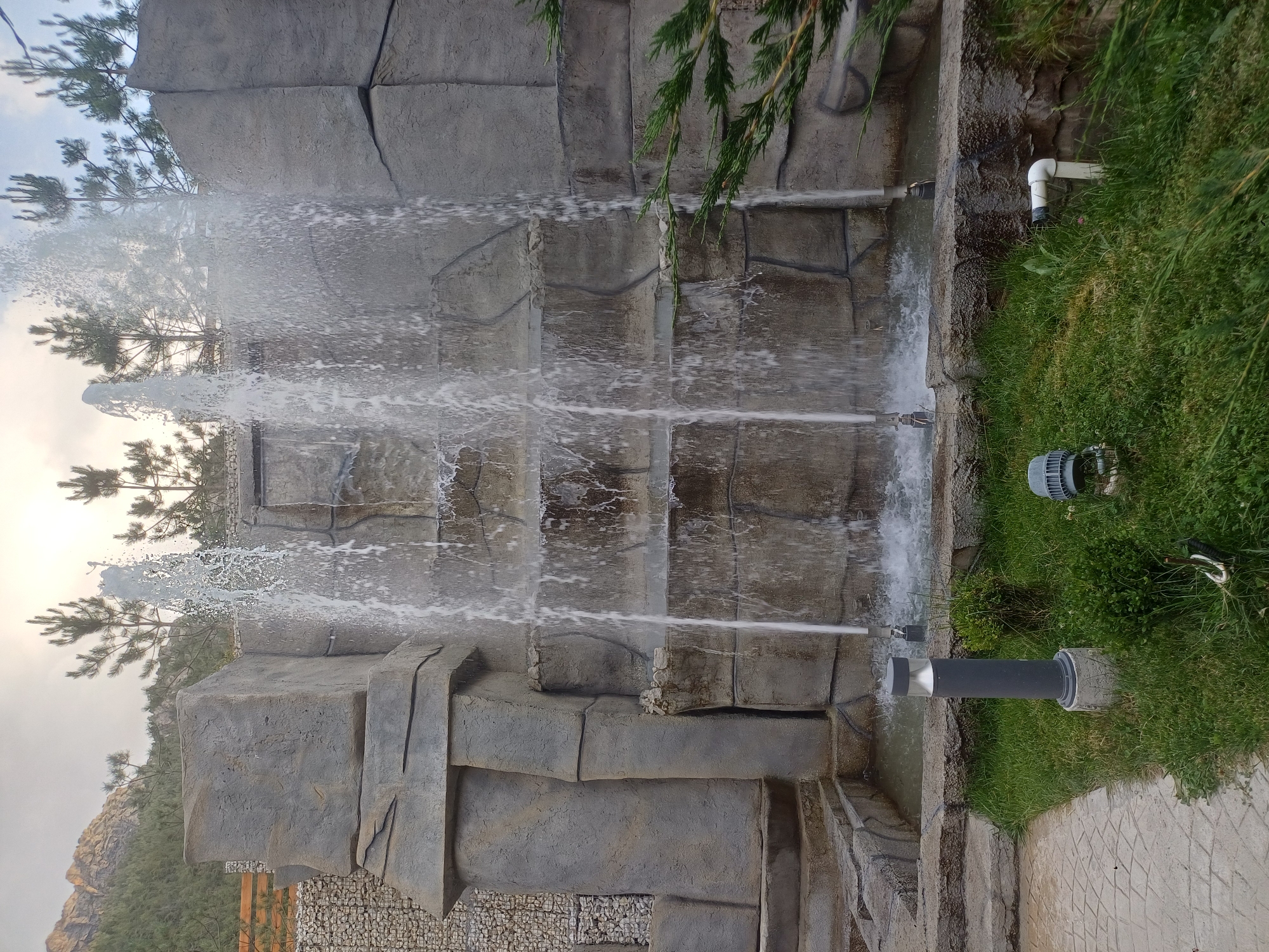 The image shows a multi-tiered artificial waterfall structure made of gray, stone-like material. Water cascades down the levels, creating a spray. In the foreground, there is a patch of green grass with several garden lights. One light is a tall, gray cylindrical lamp on a concrete base. Another, partially visible on the right, is a gray, dome-shaped light. A white pipe emerges from the ground on the right side of the frame. To the left, a paved pathway with a diamond pattern leads into the scene. In the background, pine trees are visible against a cloudy sky, with rugged, mountainous terrain in the far distance. The scene appears to be an outdoor recreational area.
