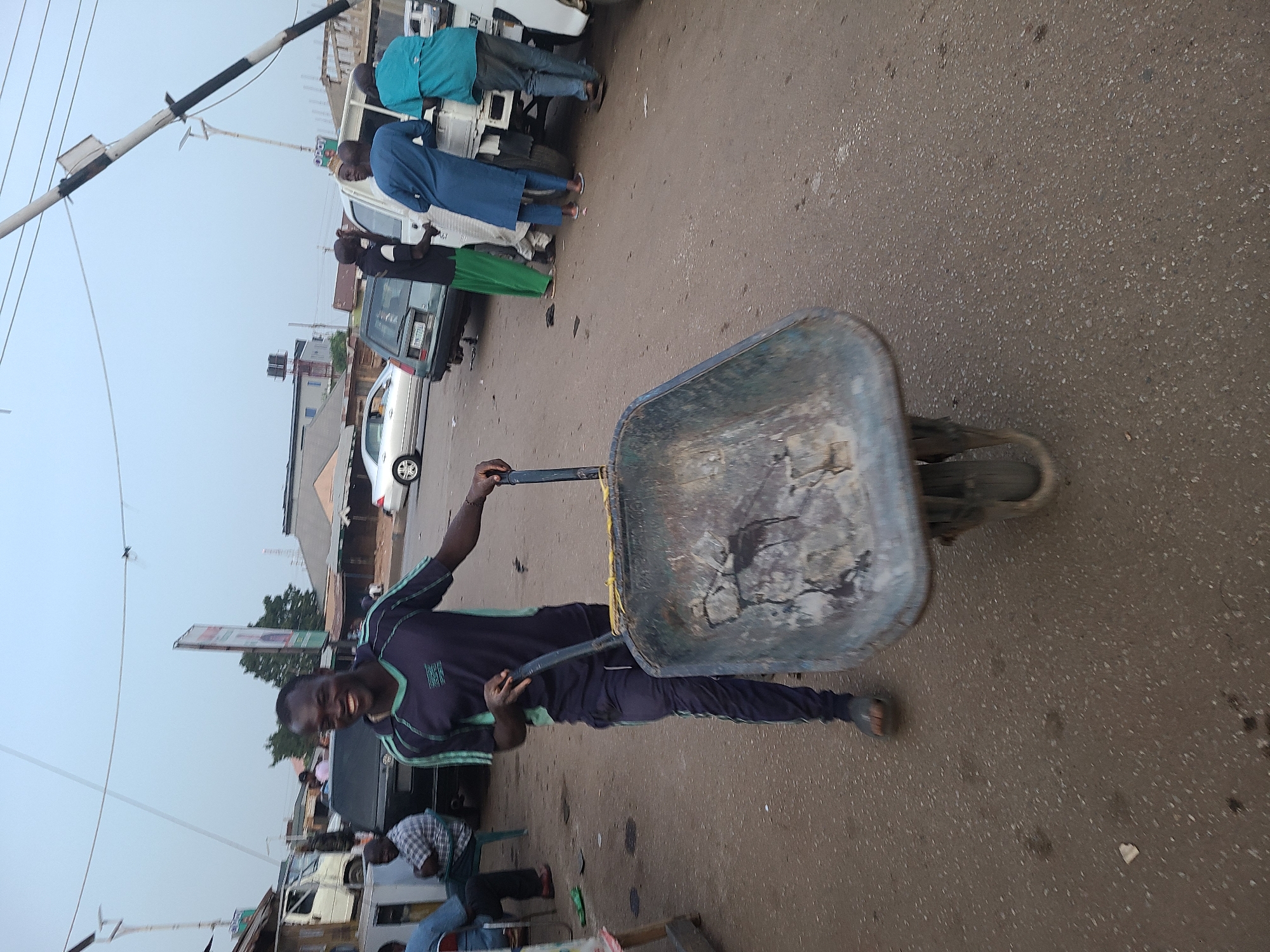 The image depicts a man in Jos, Nigeria, pushing a wheelbarrow on an asphalt street. The man wears a dark tracksuit with light green stripes and appears to be smiling. The wheelbarrow is old and contains dirt or debris. In the background, there are several people standing near vehicles, including cars and trucks. Buildings and utility poles are also visible in the background under a hazy sky.