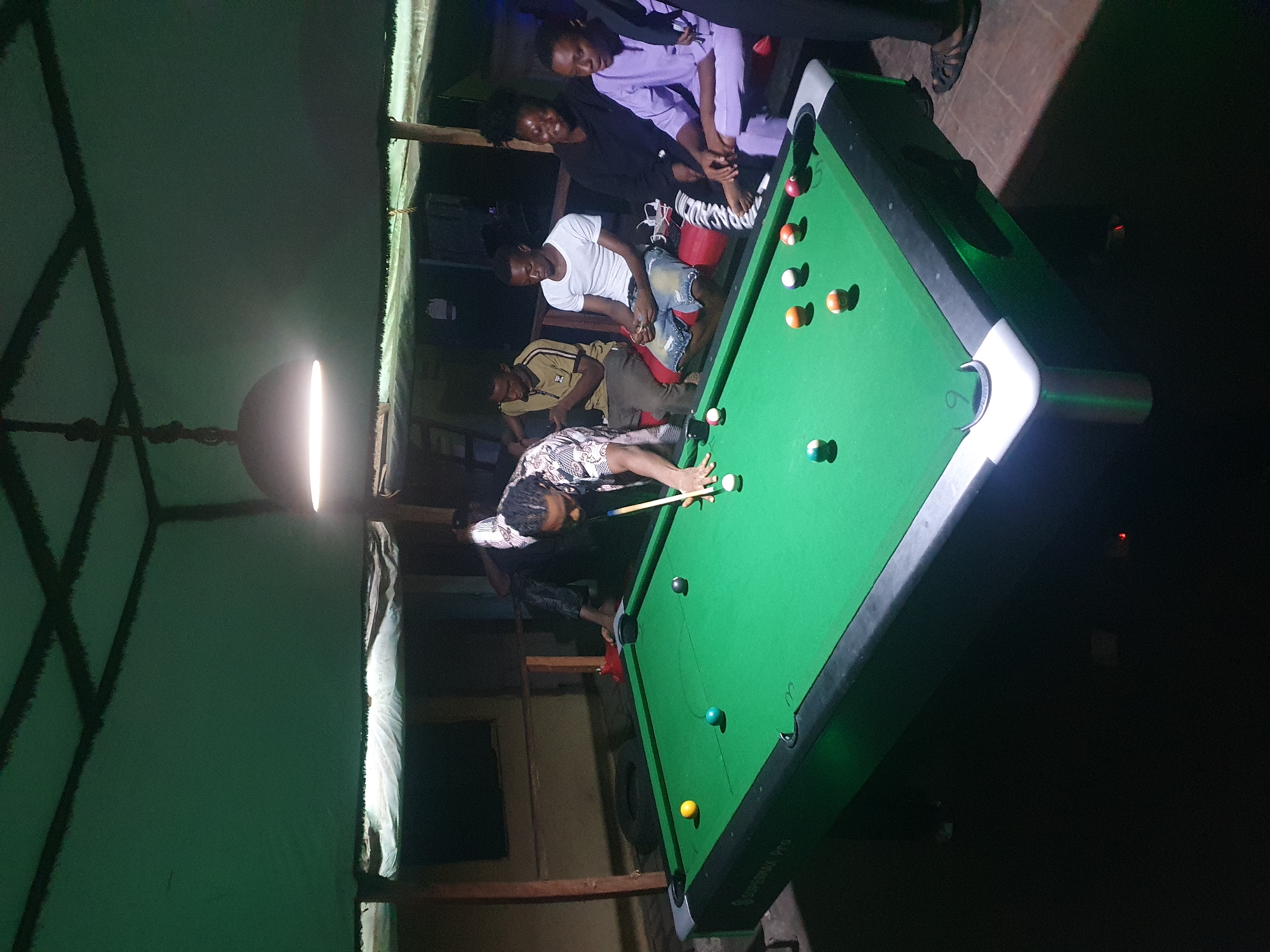 This image captures a group of people engaged in a game of pool in an indoor setting at night in Lokoja, Nigeria. The scene is dominated by a bright green pool table, centrally illuminated by a hanging light fixture directly overhead.
A person wearing a patterned shirt is actively playing, leaning over the table with a cue stick, focused on striking the cue ball amidst several scattered colored and striped pool balls. Around the table, several onlookers are gathered. Two men are seated on a bench or stool on the far side; one wears a white T-shirt and denim shorts, and the other is dressed in a striped polo shirt and dark pants. Standing behind them are two more individuals, one in a light purple outfit and another in a dark top. The atmosphere appears casual and social.
The surroundings are dark, indicating it is nighttime, with artificial light providing the main illumination. Visible in the background are wooden posts and beams, suggesting a rustic or informally constructed indoor space. A red bucket is on the floor near the pool table. The words "SUPREMA PRO" are faintly visible on the side of the pool table.