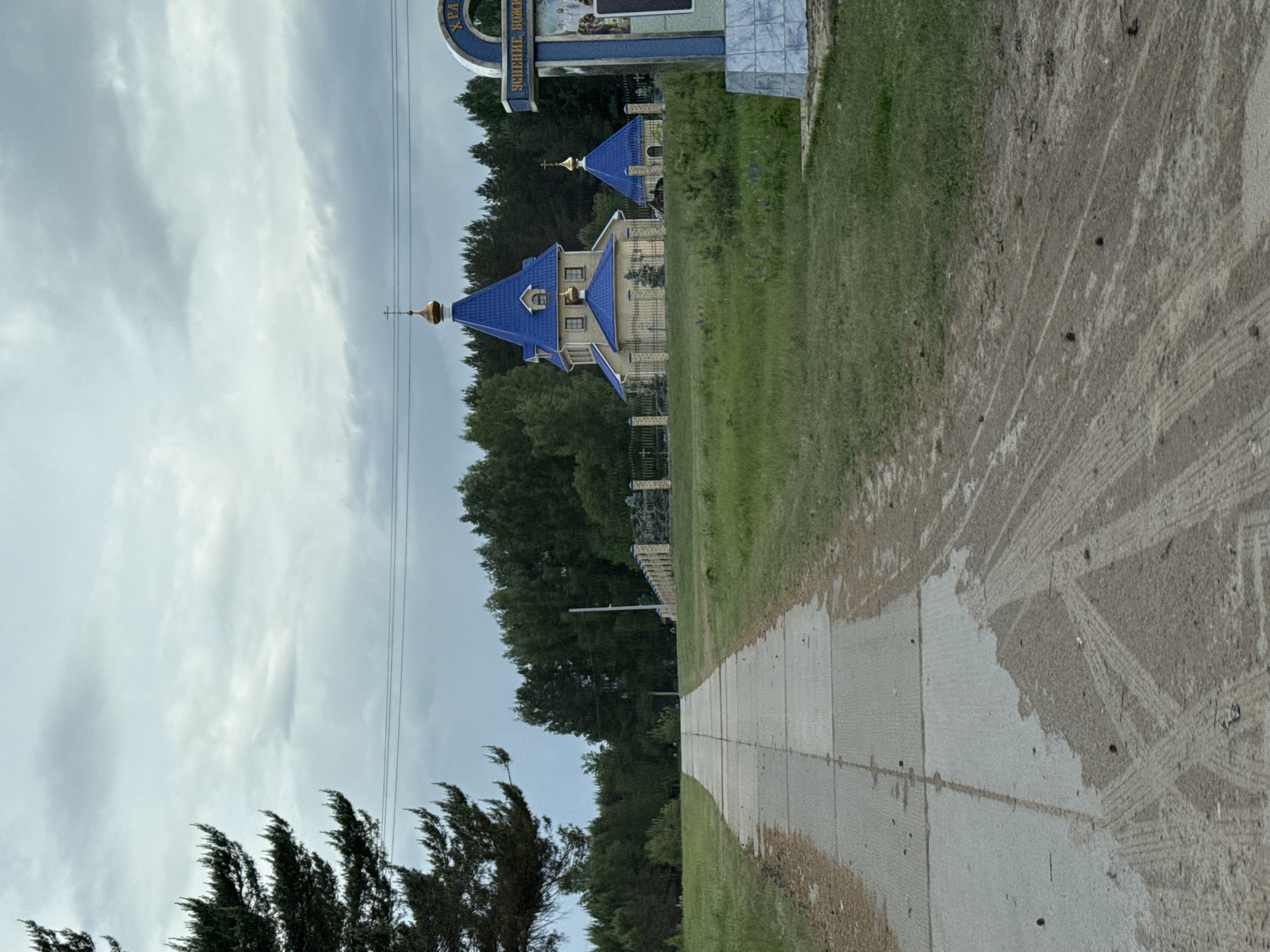 The image depicts a ground-level view of an area in the city of Chaikovsky, Russia. A paved path extends through the middle of the frame. To the right of the path is grassy terrain with tire tracks. To the left, the path is bordered by trees. Beyond the trees, a blue-roofed building with a gold cross on top can be seen. A section of fence runs along the perimeter of the property. At the top of the frame, a sign is visible. The sky is overcast.
