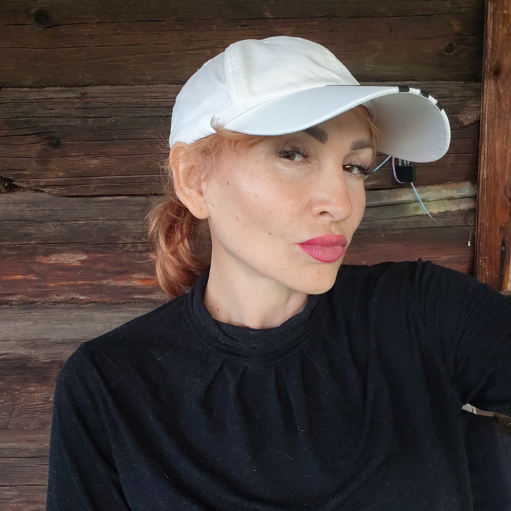 A woman is shown in a medium shot, wearing a black turtleneck sweater and a white baseball cap. The cap has a black and white striped trim under the brim. Her reddish-blonde hair is pulled back into a ponytail. She is wearing lipstick. The background appears to be a rustic, wooden wall. The photograph was taken in Chaikovsky, Russia.