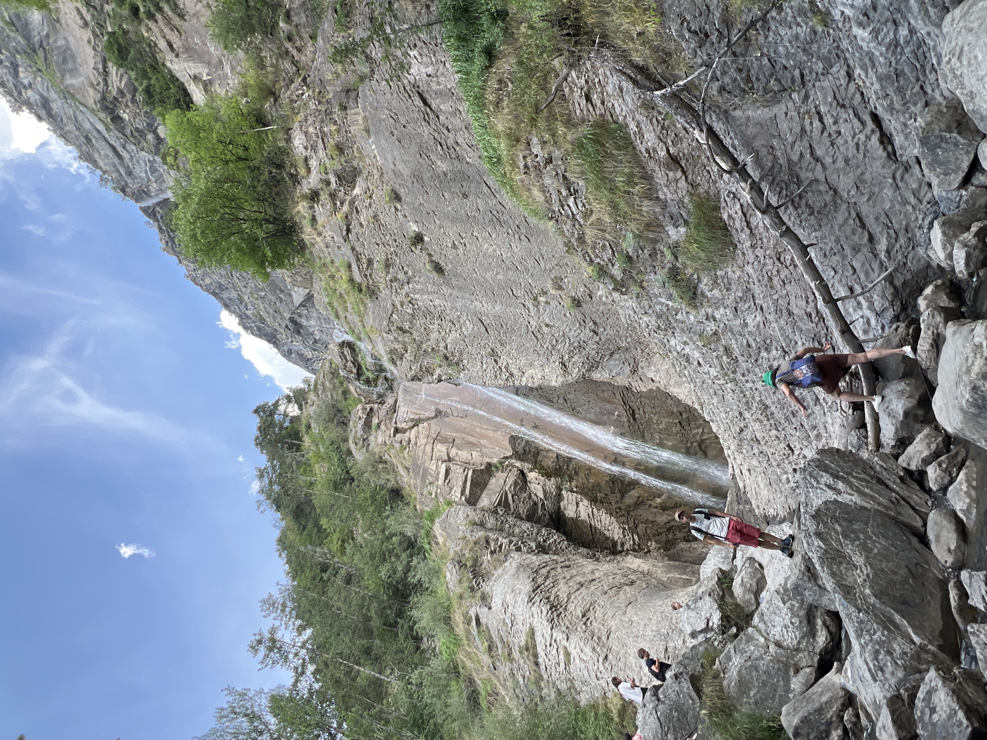 A low-angle, full shot captures a group of people hiking near a tall, narrow waterfall cascading down a steep, rocky cliff face under a bright, partially cloudy sky. The waterfall originates from the upper part of the cliff and descends a significant distance, appearing as a thin ribbon of white water against the grey rock.
In the foreground, a man stands on a large grey boulder, facing the camera. He wears a white t-shirt, red shorts, a tan baseball cap, and a backpack. To his right and slightly further down the rocky slope, a person with a green hat, brown shorts, and a backpack is climbing over rocks, facing away from the camera.
Further up and to the left, several other individuals are visible on the rocks. One person in a white t-shirt and dark shorts stands with their back to the camera, looking towards the waterfall. Another person in a black t-shirt and dark shorts is partially visible behind them. A smaller figure, possibly a child, in a light-colored shirt and shorts, is standing on a rock to the left of the man in red shorts.
The environment is rugged and natural, characterized by large, grey boulders, scree, and sparse vegetation like grass and shrubs growing on the rocky slopes. The cliff face itself is textured and shows signs of erosion. Dense green trees line the upper portion of the cliff on the left side of the image. The sky above is a vibrant blue with scattered white clouds, one of which is a small, distinct puff in the upper left quadrant. The scene is consistent with a hiking or recreational area, likely a natural gorge or mountain trail, such as those found in the vicinity of Sallanches, France, known for its mountainous terrain and natural beauty.