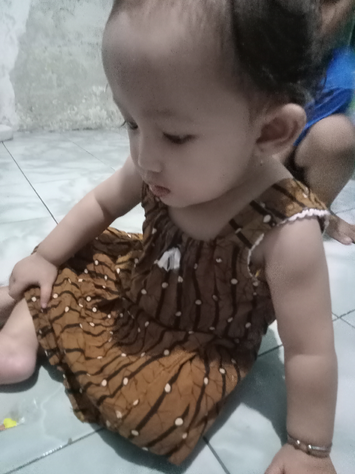 A child sits on a tiled floor, wearing a brown dress with white dots, possibly in a residential area in Bungur, Indonesia. Another individual wearing a blue shirt is partially visible behind the child. The floor tiles are light-colored with dark grout lines. The wall in the background is off-white and appears somewhat aged.