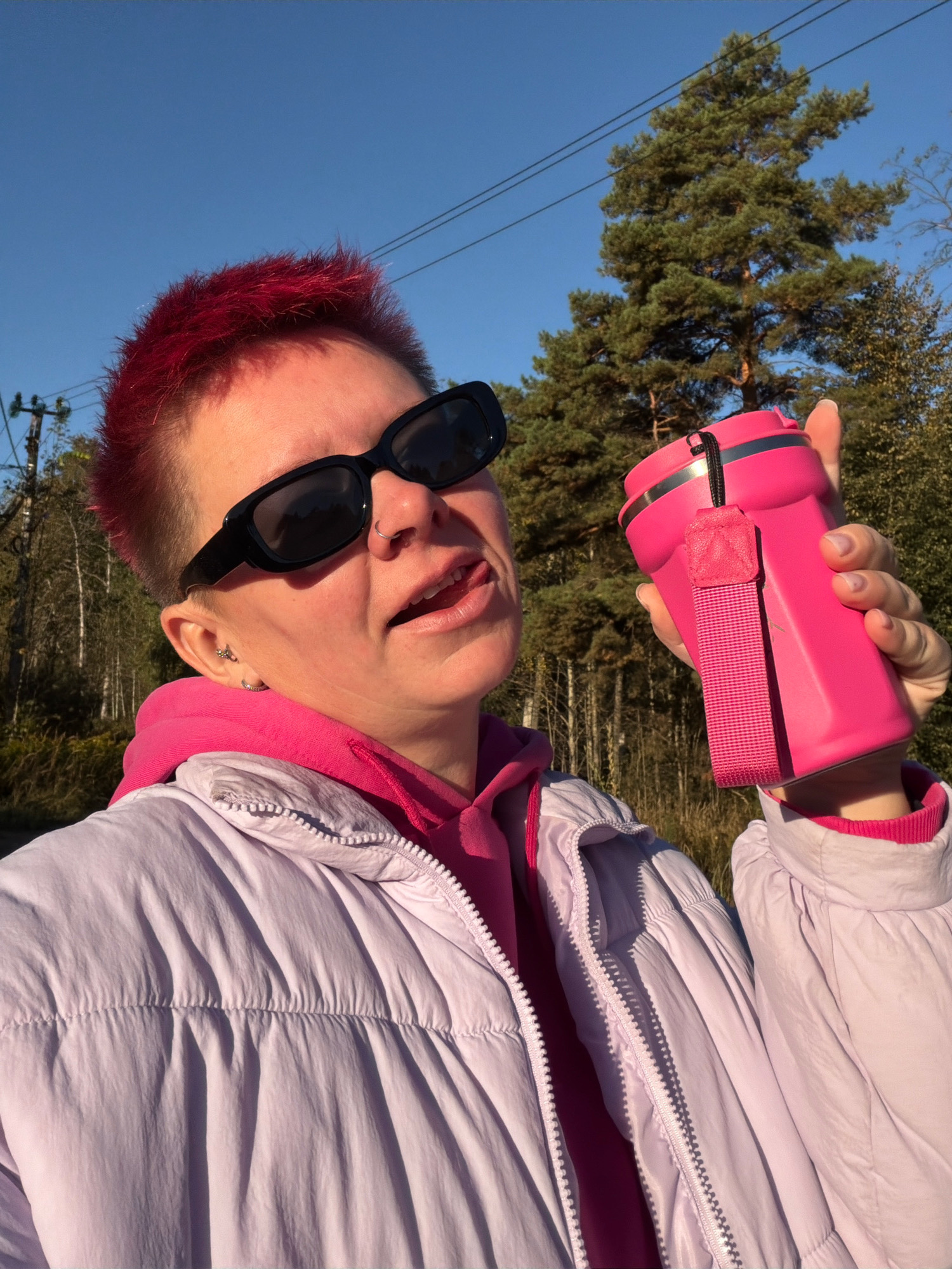 In a close-up, low-angle shot, a person with short, bright red hair, wearing black rectangular sunglasses, a pink hoodie, and a light purple puffer jacket, holds a bright pink insulated tumbler. The person is sticking their tongue out slightly, with a nose ring visible. They are outdoors, with a clear blue sky above and trees, including a tall pine, in the background. Power lines are visible against the sky. The lighting suggests it is daytime, with the sun casting shadows. The image appears to have been taken in or around Chaikovsky, Russia, given the presence of specific types of trees and electrical infrastructure commonly found in the region.