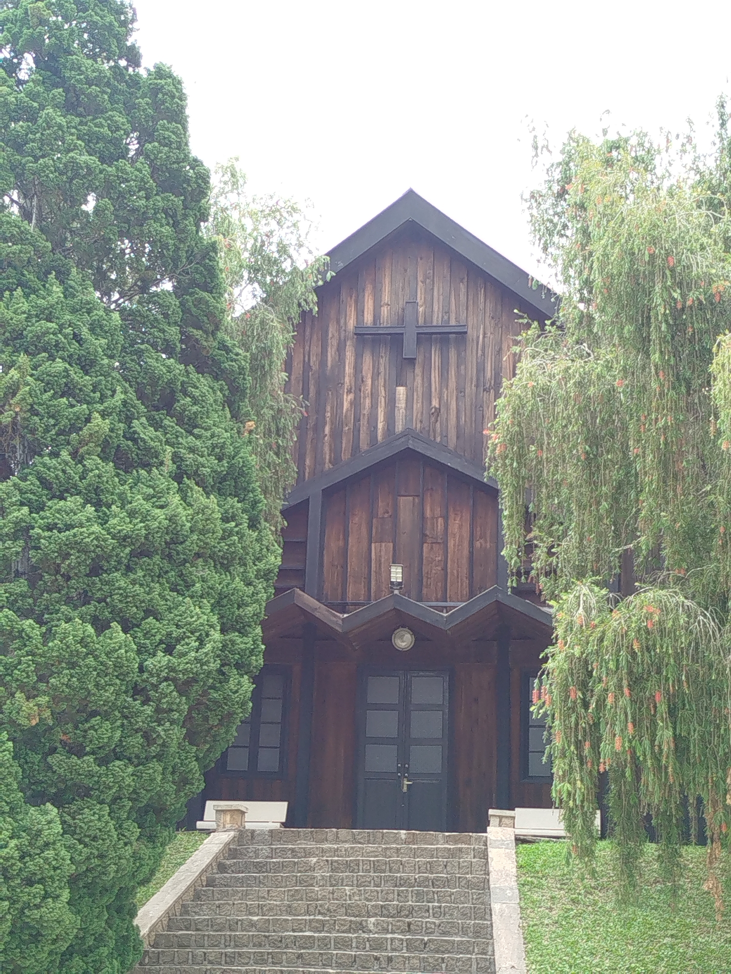 The image shows a wooden church facade in Lâm Viên, Đà Lạt, Vietnam. The church has a prominent cross on its front and a dark roof. The entrance is reached by stone steps. Trees are on either side of the church, partially obscuring the view. A weeping tree, adorned with red flowers or seed pods, is on the right.