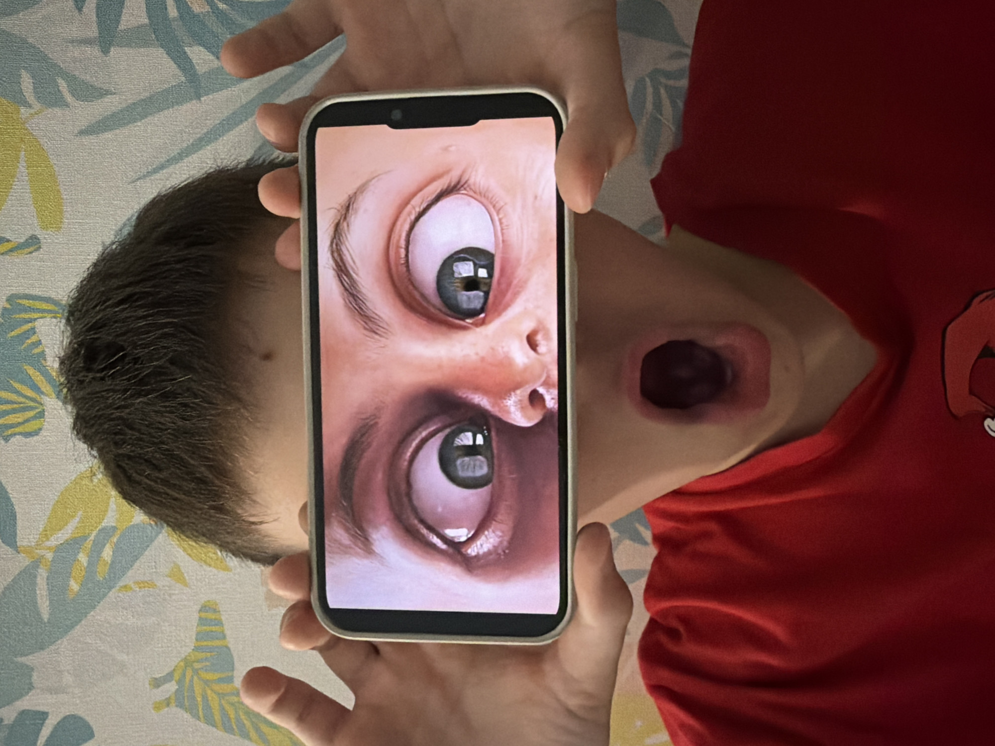 This image, taken in Votkinsk, Russia, shows a person lying down and holding a smartphone vertically with both hands. The person's head is towards the left of the frame, and they are wearing a plain red shirt or top. Their mouth is open wide, forming a dark, elongated oval shape, suggesting an exaggerated gasp, scream, or expressive action.
The smartphone's screen displays a surreal, close-up image of a human face, focusing intensely on two widely opened eyes. The upper eye is angled slightly upwards, while the lower eye is angled downwards, both featuring prominent dark pupils with noticeable reflections of light sources. The skin around the eyes is light-toned, and dark eyebrows and eyelashes are visible, contributing to an expression of intense surprise or alarm on the screen.
The hands holding the phone are relatively small, suggesting a child or young adult. The person's short, dark hair is visible on the left side of their head. The background consists of a light-colored fabric, likely bedding or a sofa, decorated with a repeating pattern of stylized blue and yellow leaves or foliage. The lighting appears bright and even, illuminating the scene clearly. No visible text is present in the image. The composition creates a playful yet slightly unsettling effect, combining the digital image's intensity with the physical person's exaggerated expression.