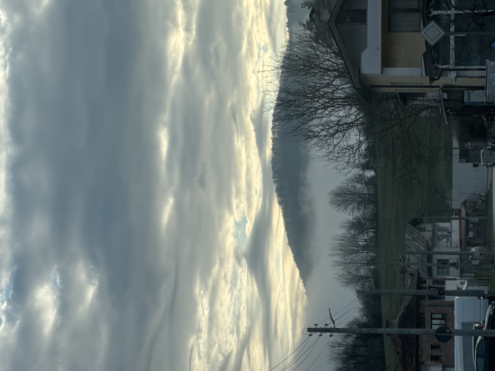 The image depicts a scene near Smilyan, Bulgaria. The sky is mostly covered with striated clouds. A mountain range runs vertically through the mid-ground, partially obscured by mist or fog. To the right, there is a mixture of vegetation, including leafless trees and grassy fields. Closer to the right edge of the image are buildings, including houses and commercial structures, along with street signs and utility poles with wires. Some vehicles are parked on the right side of the frame.