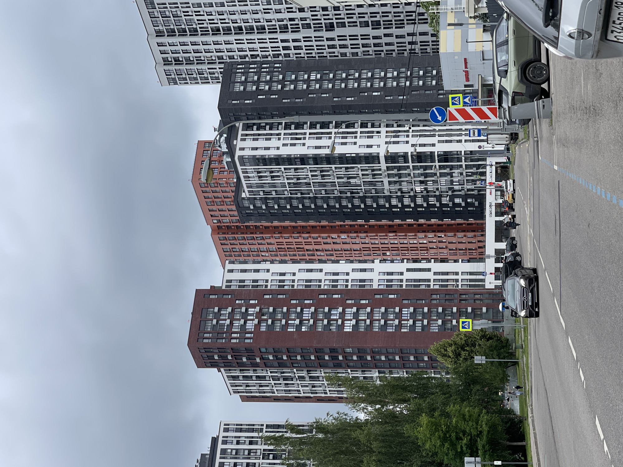 A low-angle shot shows a line of modern, multi-story apartment buildings with facades of white, black, and reddish-brown brick, interspersed with numerous windows. In the foreground, a paved road runs alongside the buildings, with a car in motion. Traffic signs are visible on the side of the road, including a blue circular sign with a white arrow indicating a turn and a diagonal striped red and white sign. The sky is overcast, suggesting a cloudy day. A few trees are visible between the road and the buildings. The scene appears to be a residential urban area. No people are clearly discernible. The overall impression is one of contemporary city architecture and daily life on a cloudy day in Moscow, Russia.