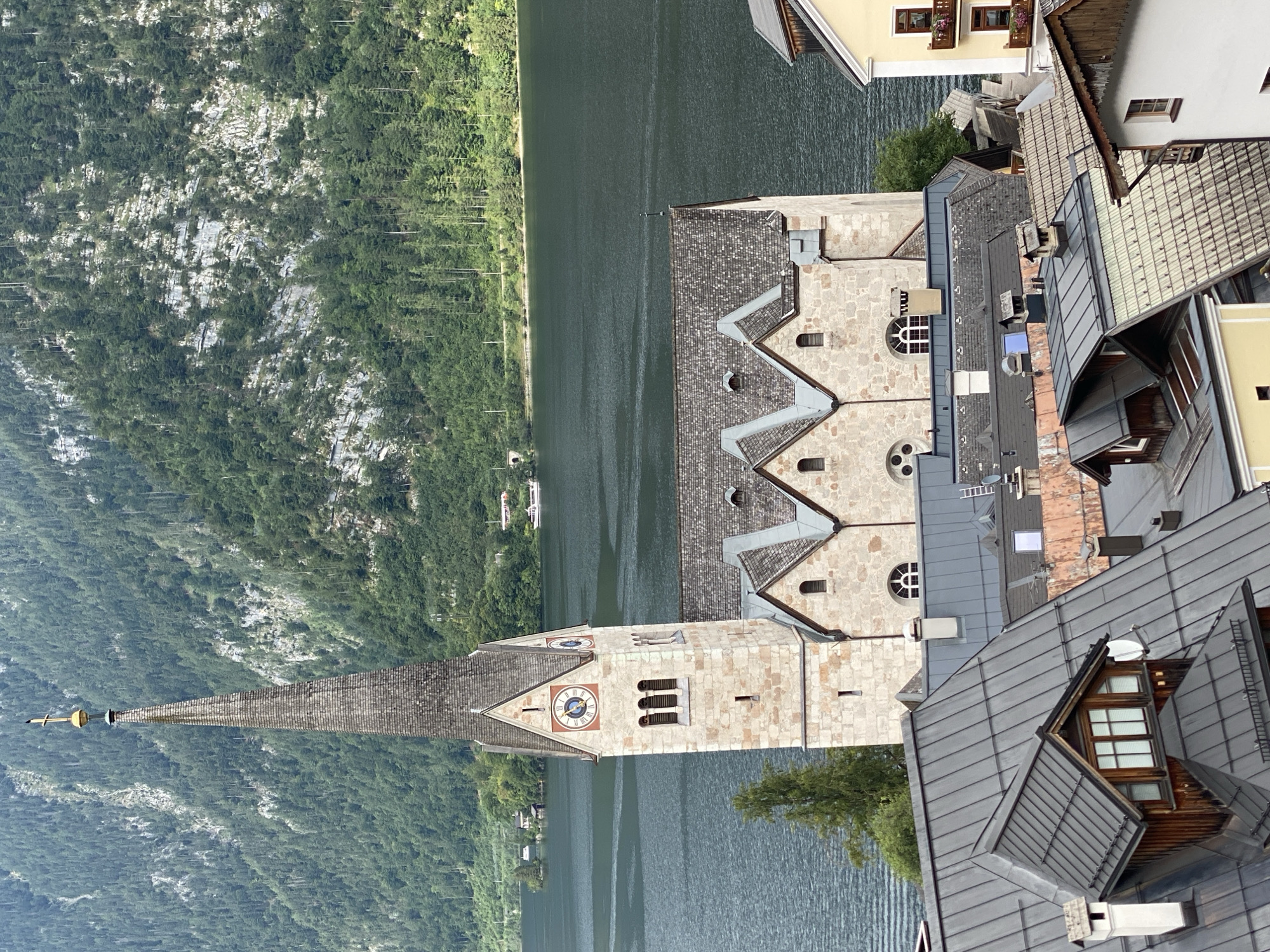 This is a high-angle, exterior, eye-level shot of the town of Hallstatt, Austria, in daylight, with a cloudy sky. The main subject is the Church of the Assumption, a Gothic Revival church with a tall, slender steeple topped by a cross. The church is constructed of light-colored stone, with a grey, shingled roof. The steeple has a clock face with Roman numerals.
In the foreground, several buildings with grey, pitched roofs are visible, some featuring dormer windows and chimneys. These buildings are closely packed, typical of a European town.
Beyond the church and foreground buildings, a large, dark body of water, likely Hallstätter See, stretches across the midground. A white boat is visible on the water.
In the background, a steep, forested mountain rises dramatically, covered in dense green trees. The mountain exhibits exposed rock formations and striations.
The scene depicts a serene, picturesque view of Hallstatt, highlighting its unique architecture nestled between the lake and the mountains. There are no people or animals clearly visible in the image.