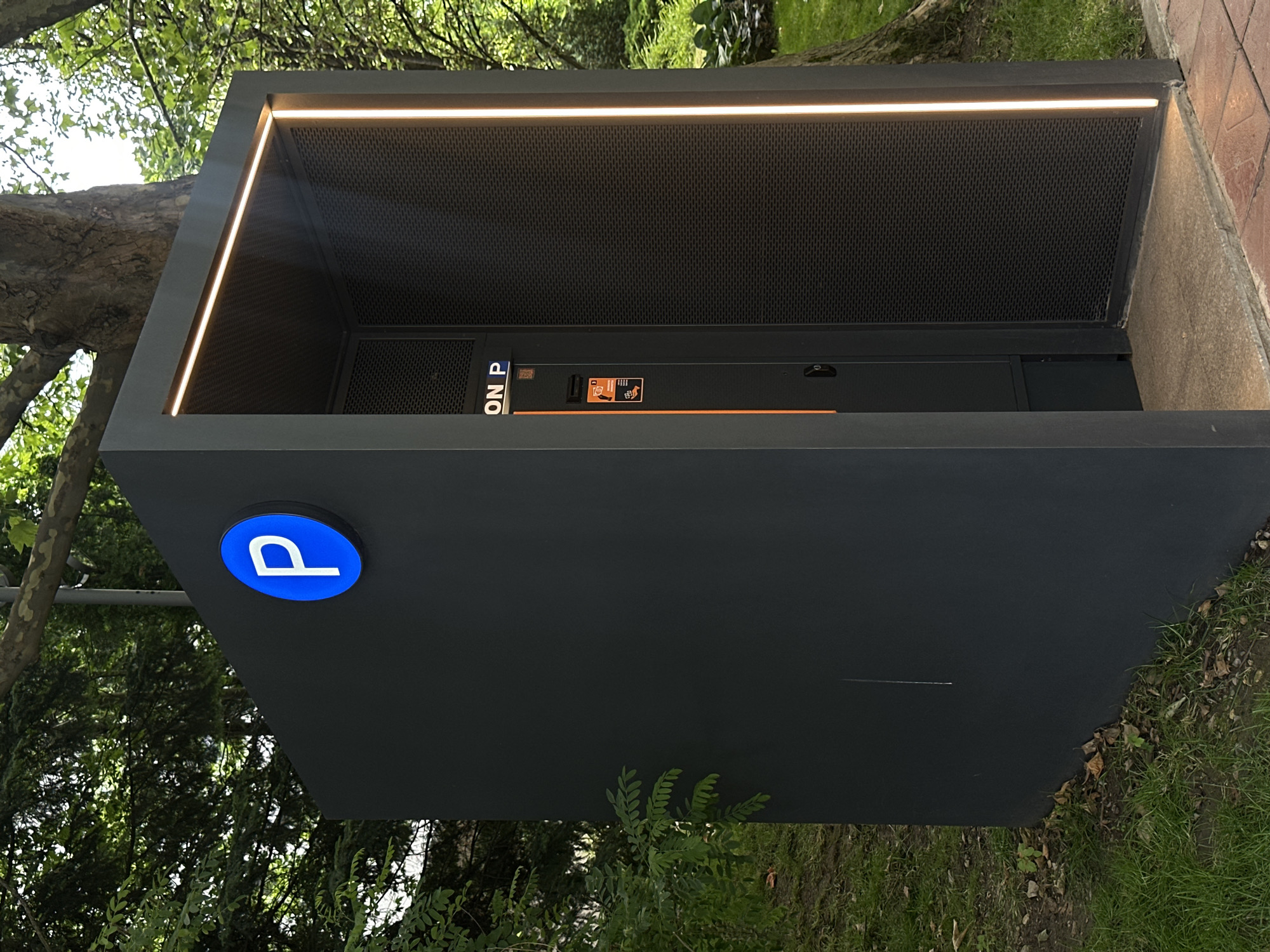 A dark gray, modern, kiosk-like structure with a blue circular sign displaying a white "P" is situated outdoors. The structure is illuminated by subtle, warm-toned LED strips along its top and side edges. Inside the open front of the structure, a black panel is visible with an orange vertical stripe. This panel has a display that reads "ON P" and a slot that appears to be for payment or ticket dispensing, along with various instructional icons. The kiosk is surrounded by greenery, with trees and grass visible in the background. The ground in front of the kiosk is paved with interlocking bricks and a stone slab. Based on the context provided by the user, this scene is located in Sofia, Bulgaria.