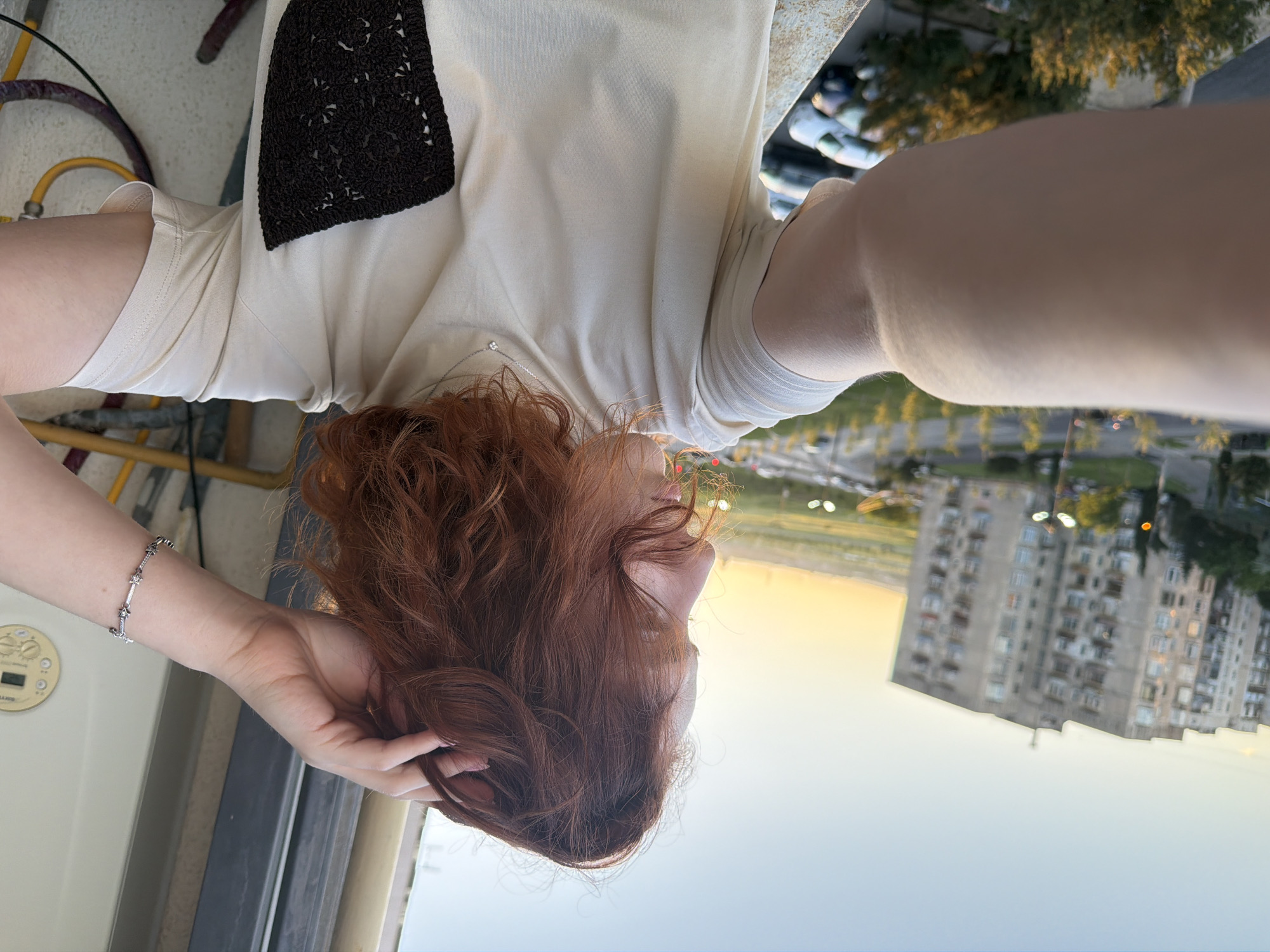 A red-haired person is positioned near a window or balcony, with a view of Tbilisi, Georgia in the background. The building's architecture suggests a residential area. The person is wearing a light-colored shirt with a dark, possibly crocheted, patch on the shoulder. A silver bracelet is visible on their wrist. The time of day appears to be late afternoon or early evening, with the sun setting and casting a warm light across the city. A road with vehicles is present in the background.
