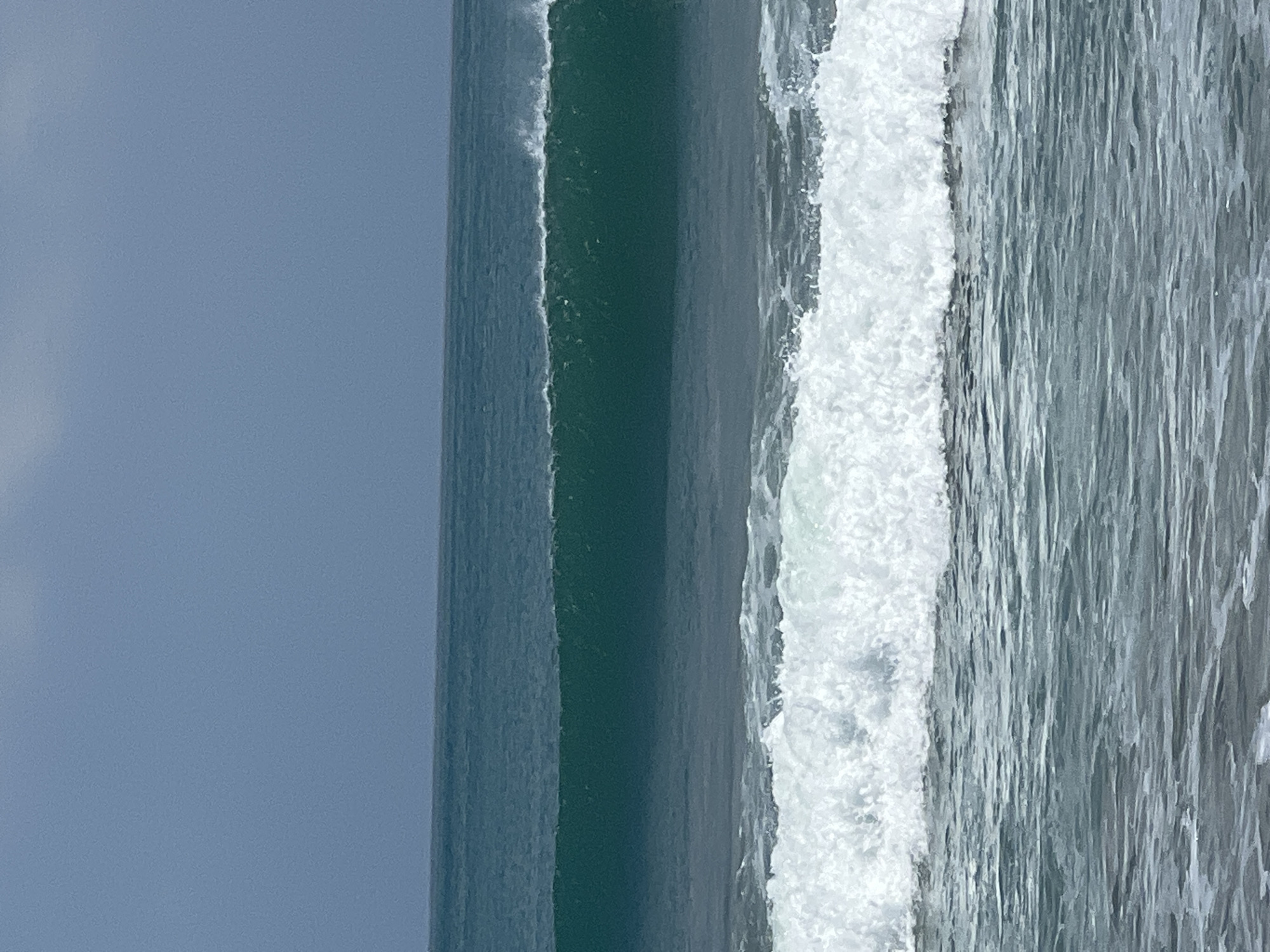 The image shows a close-up view of ocean waves on a cloudy day. The water is a deep teal color, with a large wave breaking in the foreground, creating white foam. In the background, a larger, unbroken wave is visible, its crest just beginning to curl. The sky is a uniform gray. The scene is characteristic of Kuta, Bali, Indonesia, known for its surf beaches.